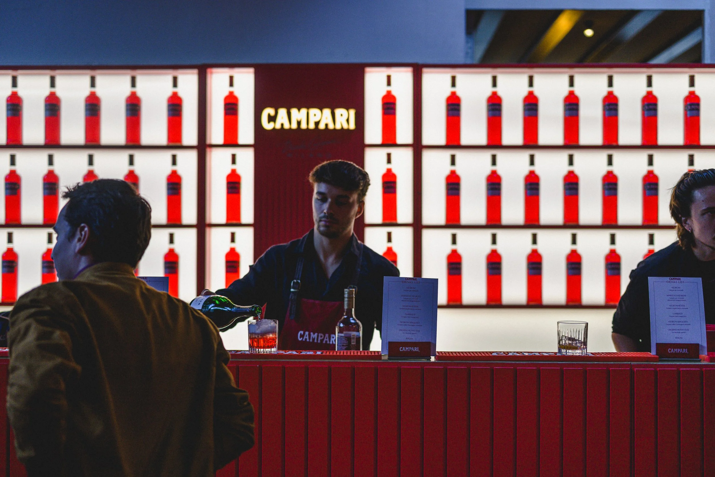 A bartender pouring a drink at a bar with a bright red backdrop featuring multiple Campari bottles and the Campari logo, while a customer with dark hair and a brown jacket stands in front of the bar.
