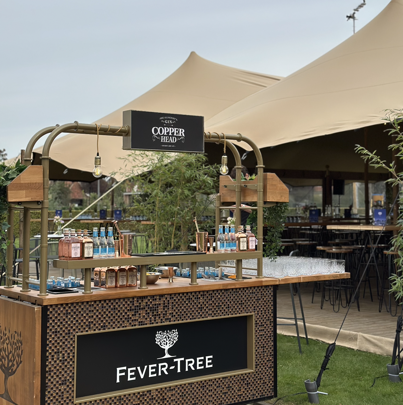 Outdoor Fever-Tree gin and tonic bar booth with Copper Head branding, various bottles, and glasses under a canopy at a festival or event.