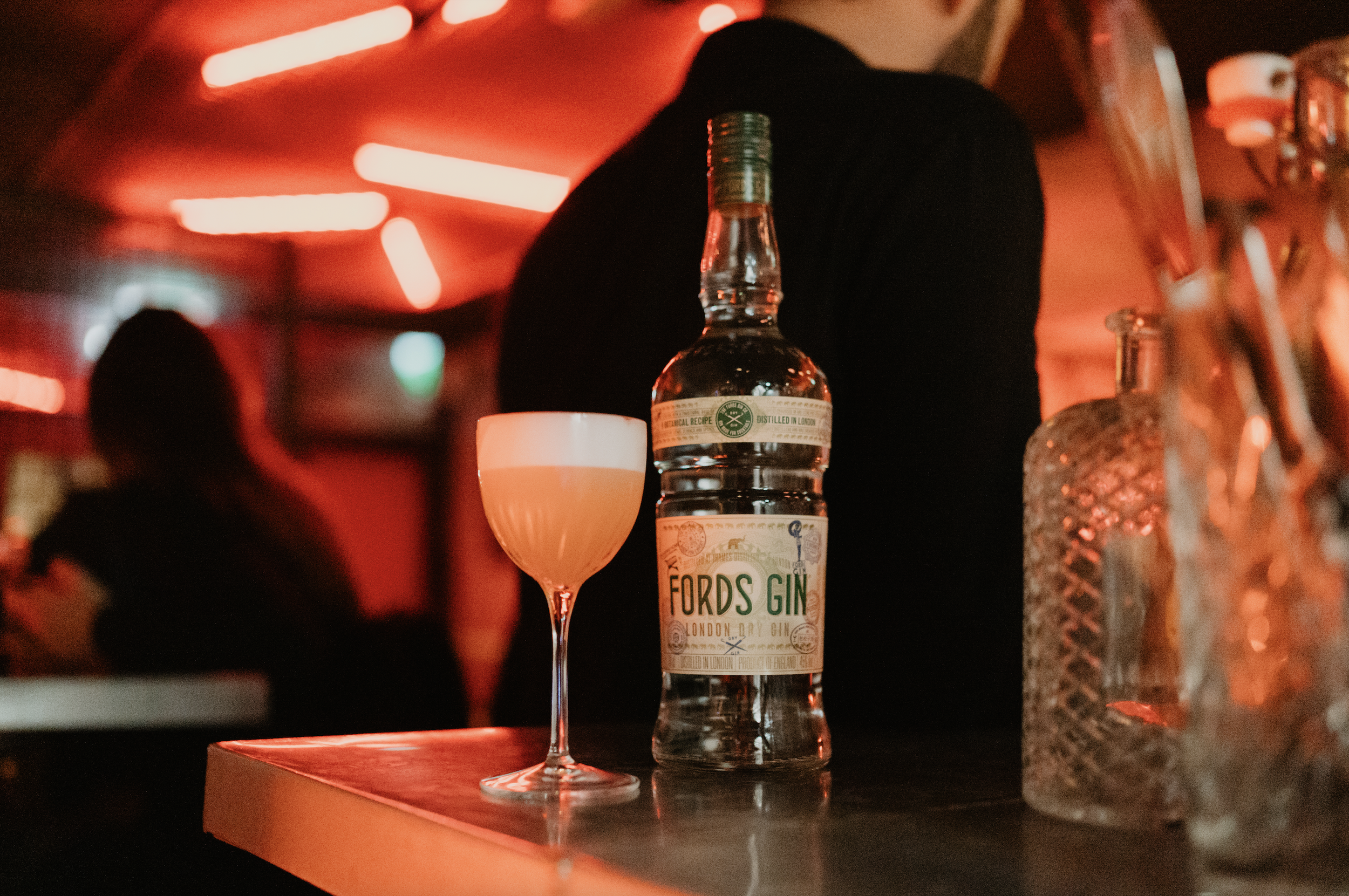 A clear bottle of Fords Gin and a cocktail glass with a light-colored drink on a bar counter. The background is dimly lit with red neon lights, and a person dressed in black is partially visible.