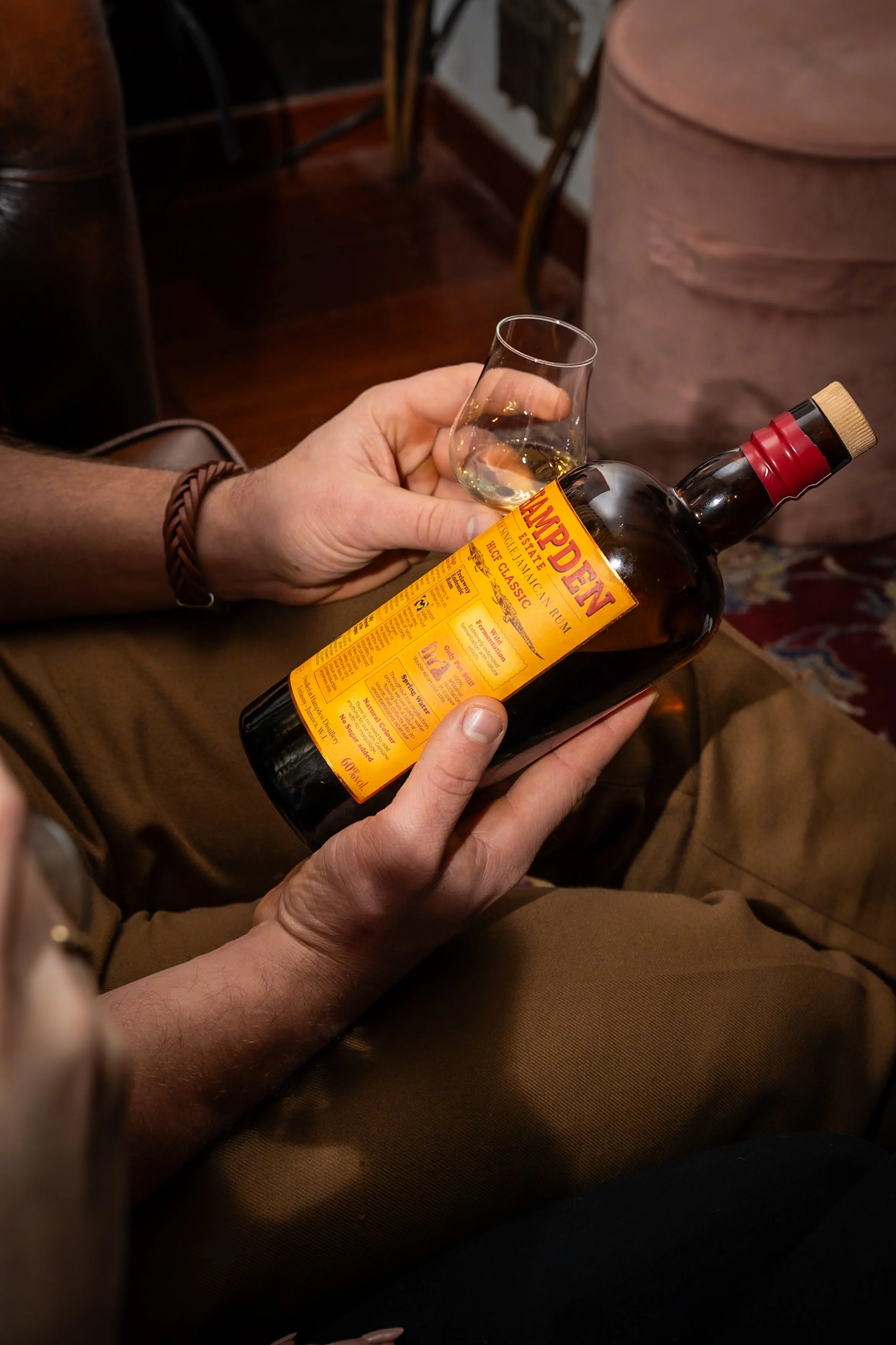 Person holding a bottle of Hampden rum and a glass with some rum inside, sitting indoors in a cozy setting.