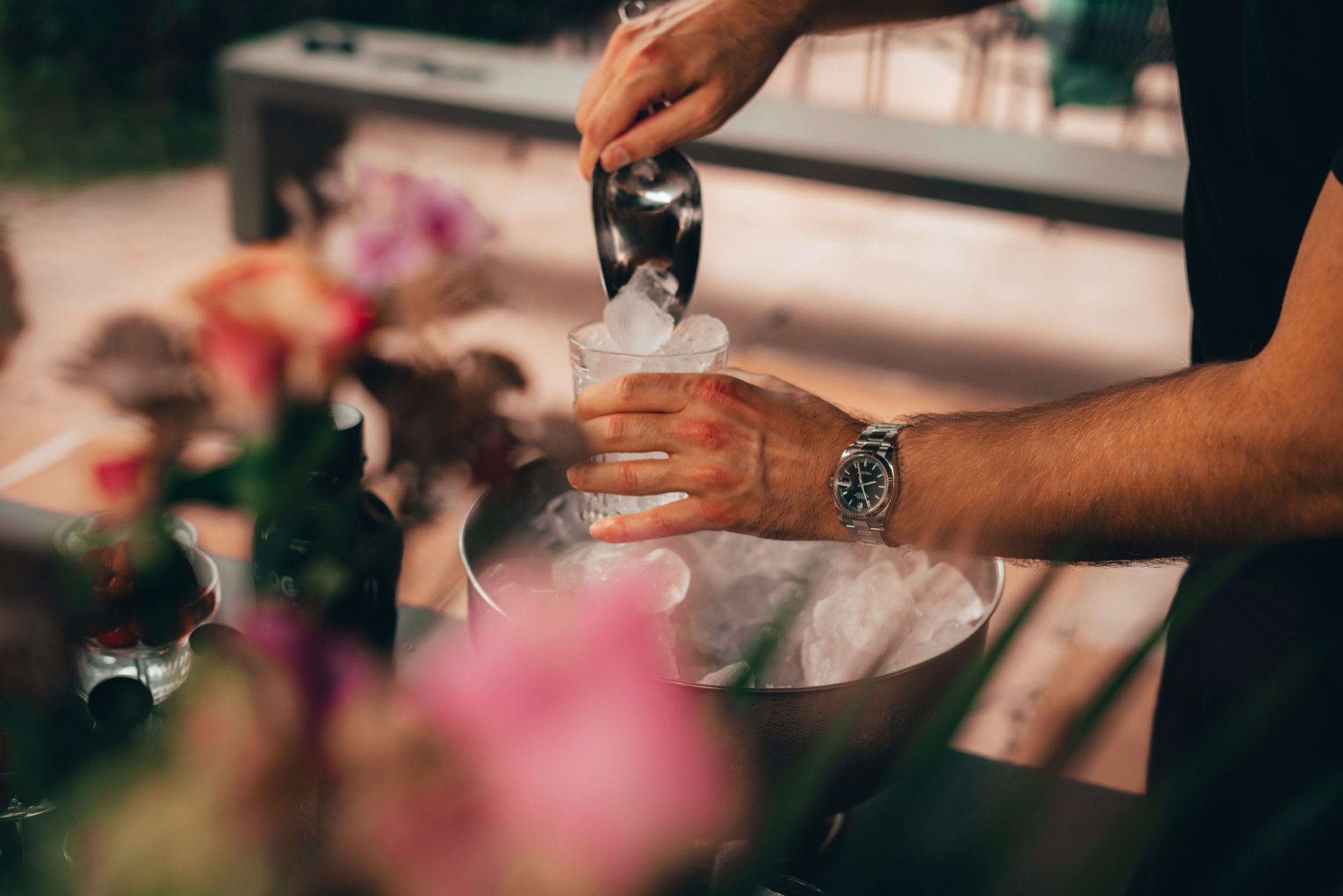 Person pouring ice into a glass with a metal ice scoop, wearing a wristwatch.