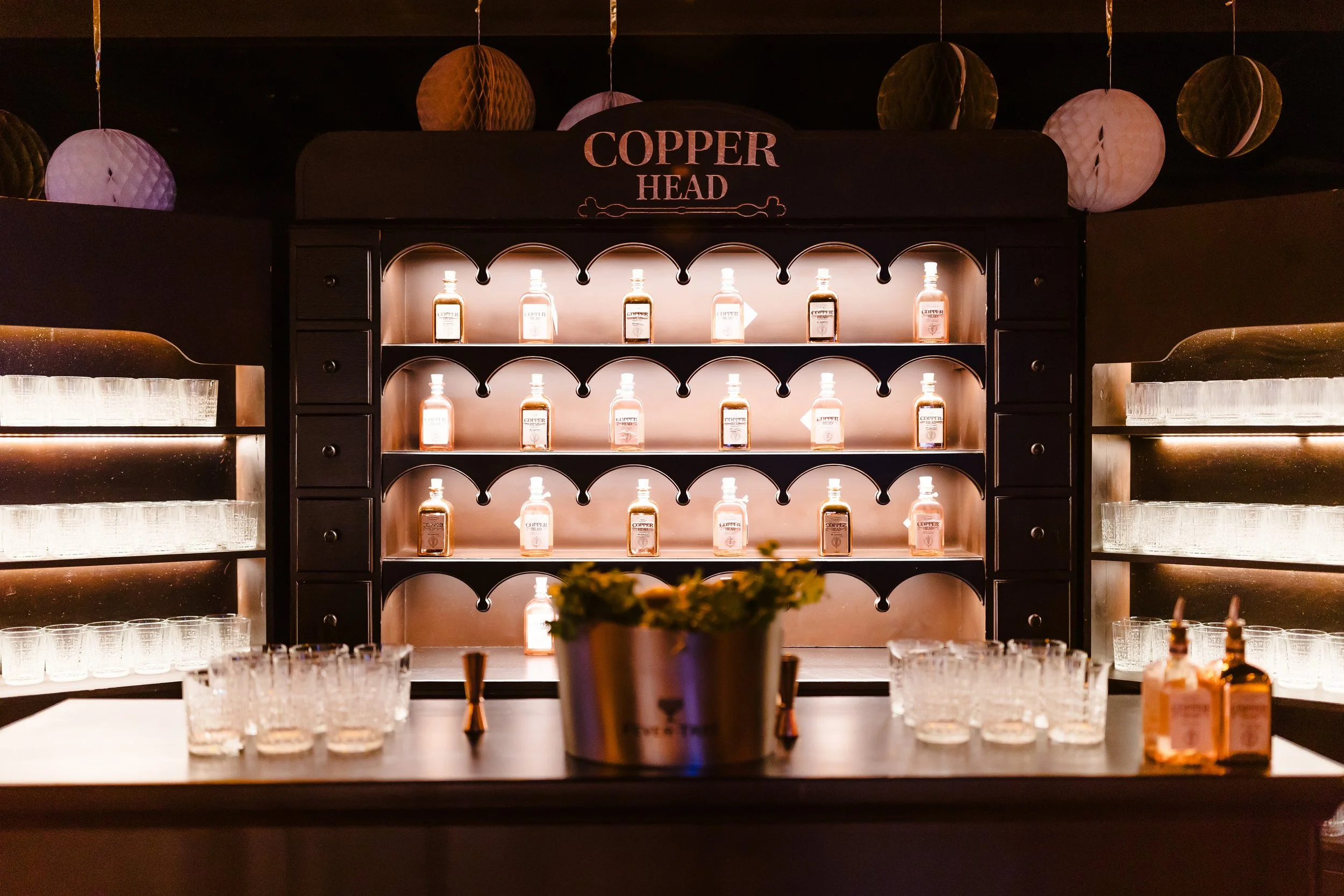 A display of Copper Head gin bottles arranged on illuminated shelves with glasses and bottles on the counter in front and decorative paper lanterns hanging from the ceiling.