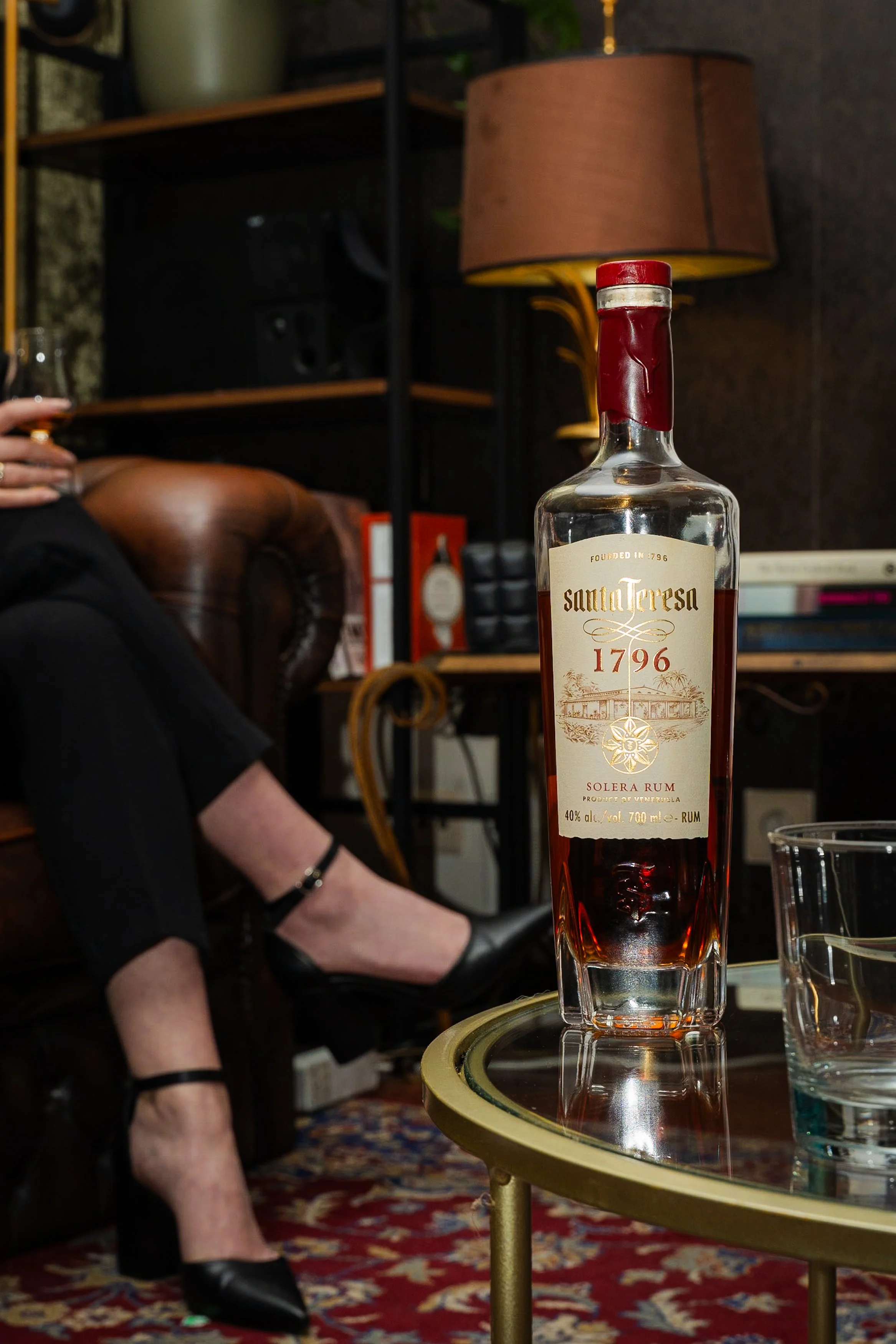 A bottle of Santa Teresa 1796 Solera Rum on a glass table next to a jar of water, with a person sitting on a leather armchair holding a glass of drink in their hand, wearing black shoes and pants, in a cozy living room setting.