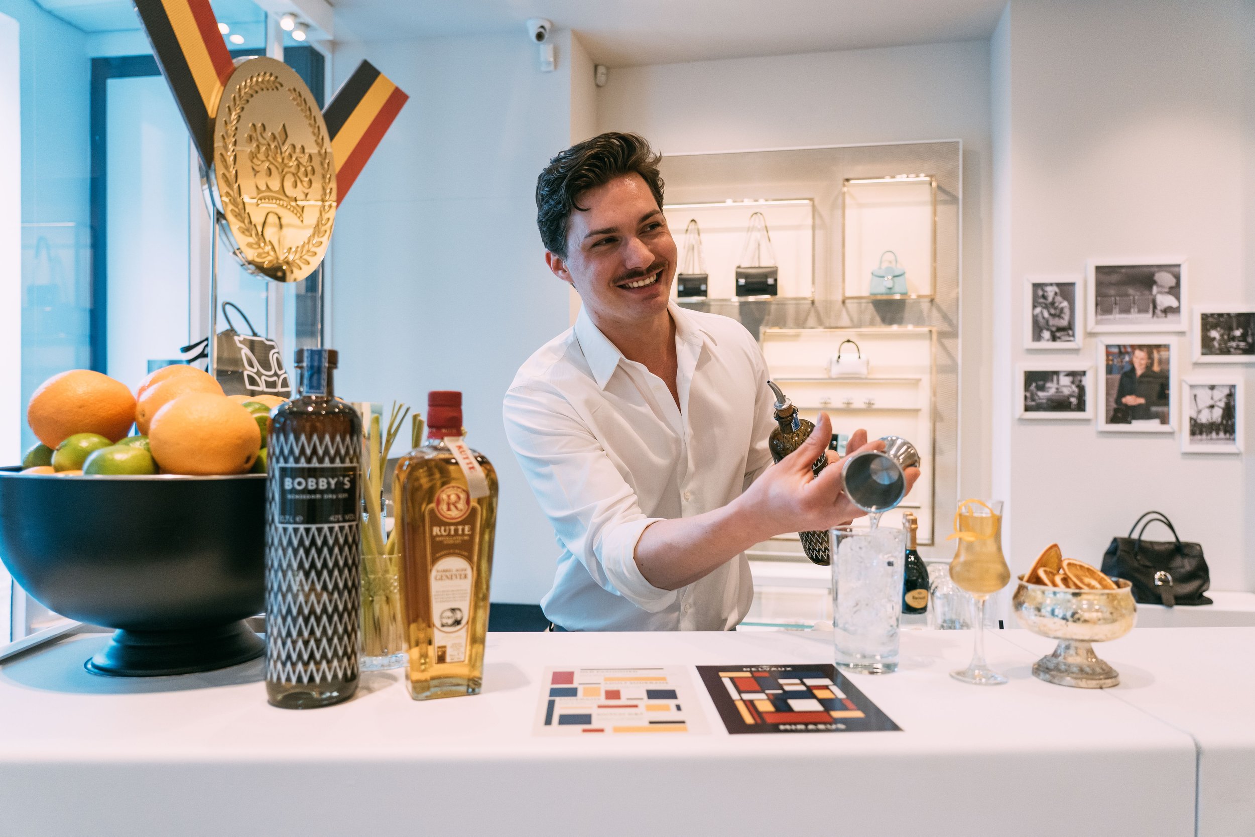 A smiling bartender in a white shirt pours a drink from a cocktail shaker into a glass at a bar counter with bottles, fruits, and bar tools, in a stylish shop or cafe setting.