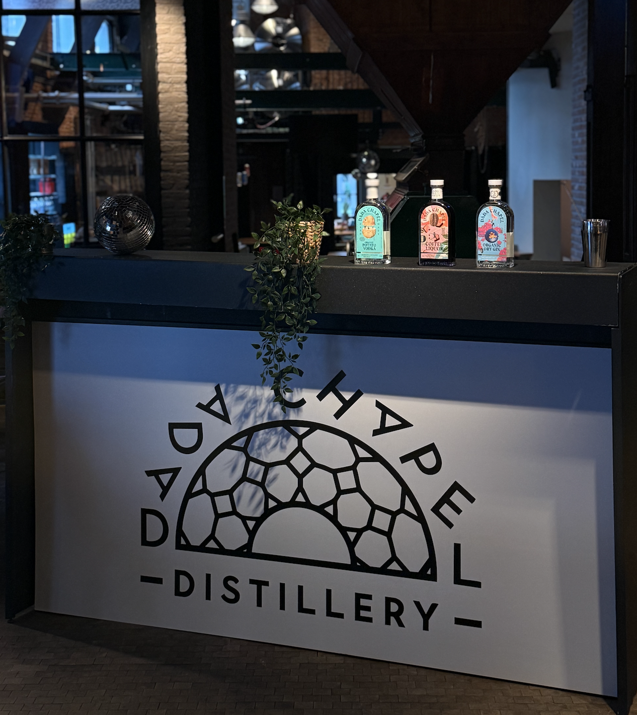 A bar counter with three bottles of gin, a shot glass, and a cocktail shaker. The front of the counter displays a sign with the logo and name 'DADA CHAPEL DISTILLERY' with a geometric sun design.