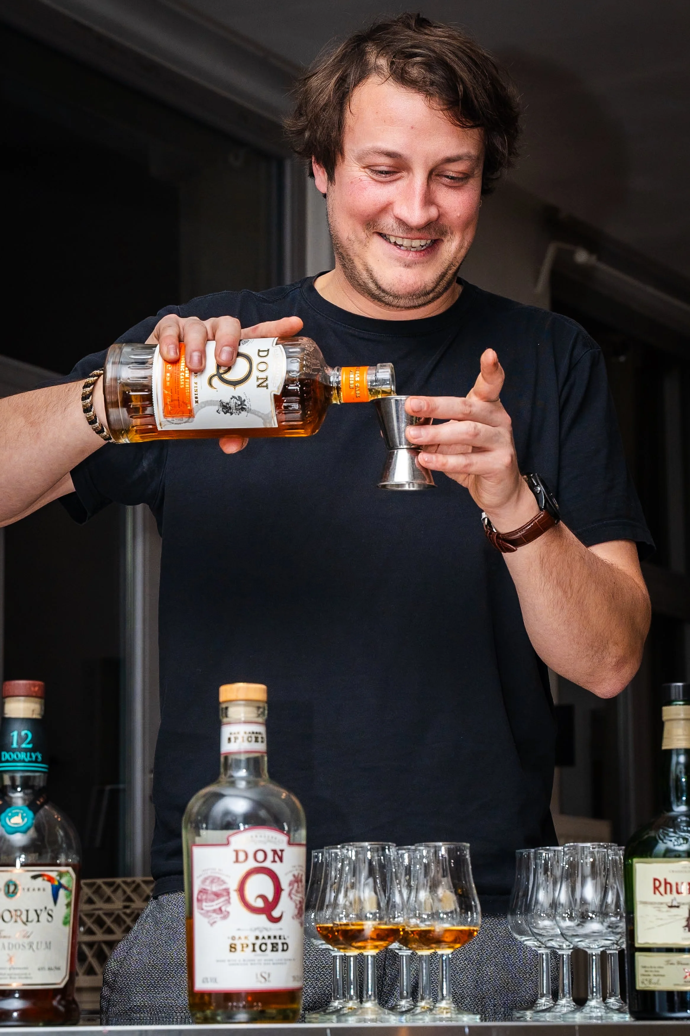 A man pouring whiskey into a glass from a bottle labeled 'Don Q' with other bottles and glasses on the table in front of him.