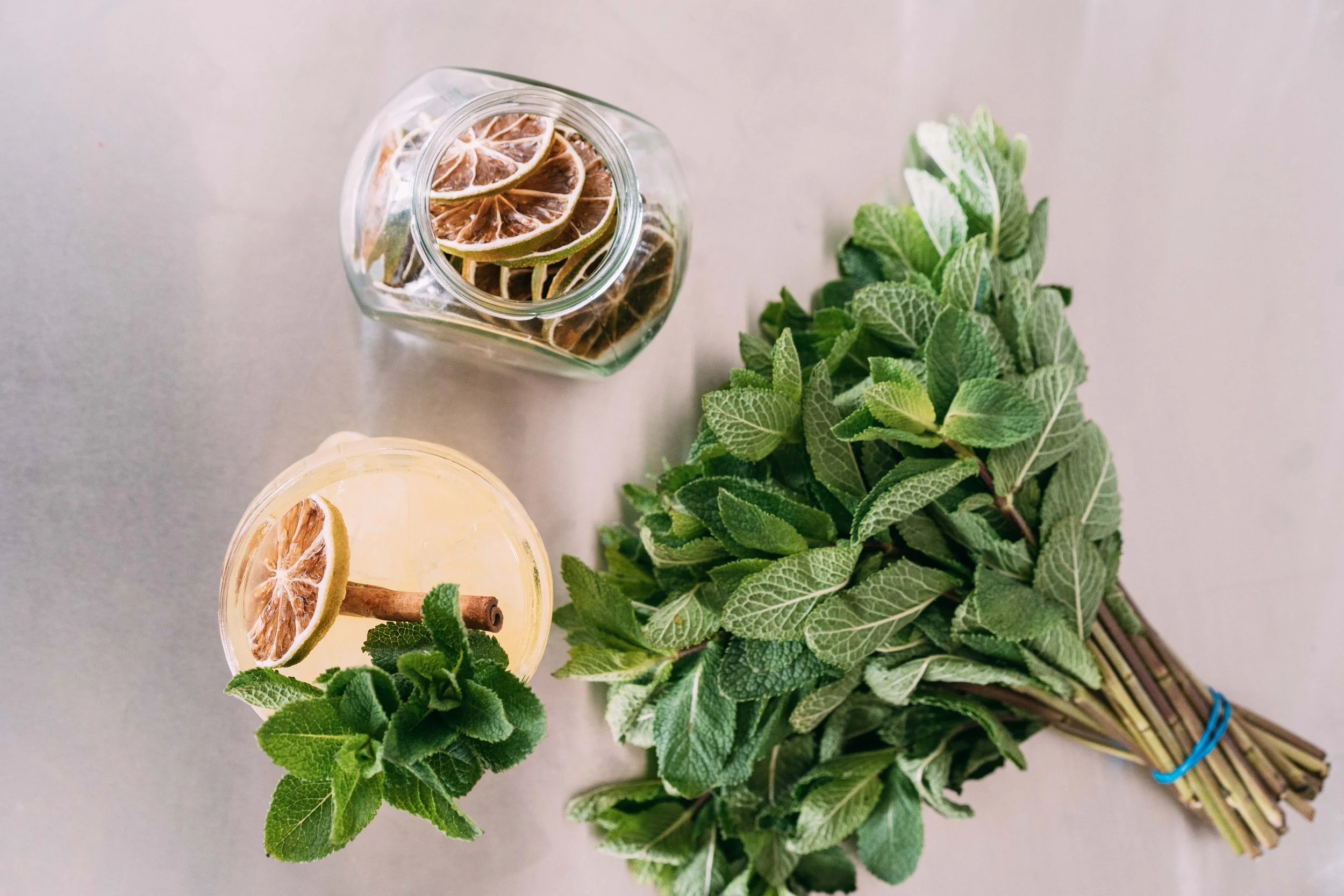 A bundle of fresh green mint leaves, a glass of lemonade with a lemon slice and cinnamon stick, and a jar of dried lemon slices on a light gray surface.
