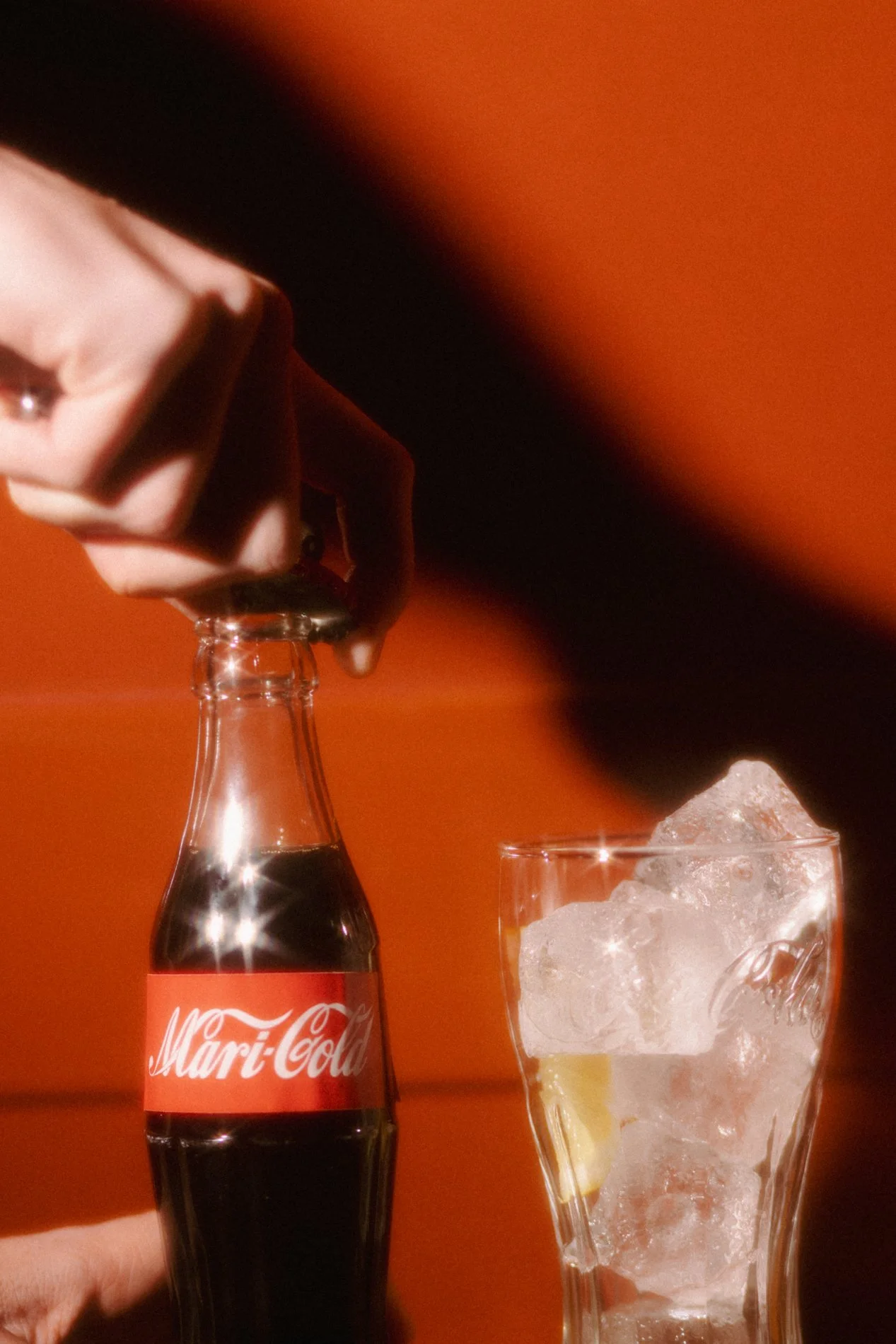 A hand holding a bottle of Marigold soda pouring into a glass filled with ice and a lemon wedge, against an orange background.