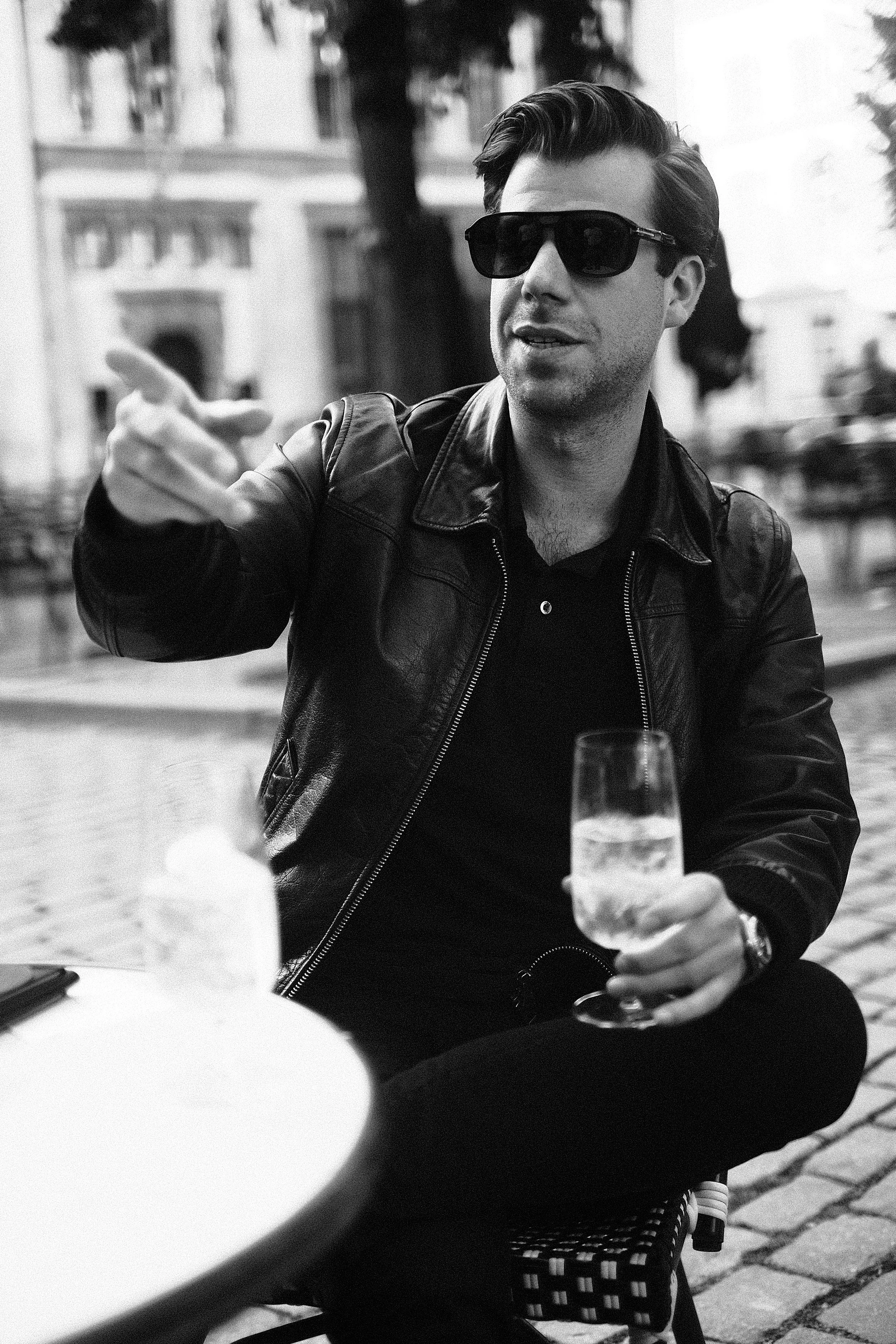 A man wearing sunglasses and a leather jacket sitting outdoors, holding a glass of champagne, with a woman in the foreground and buildings in the background.