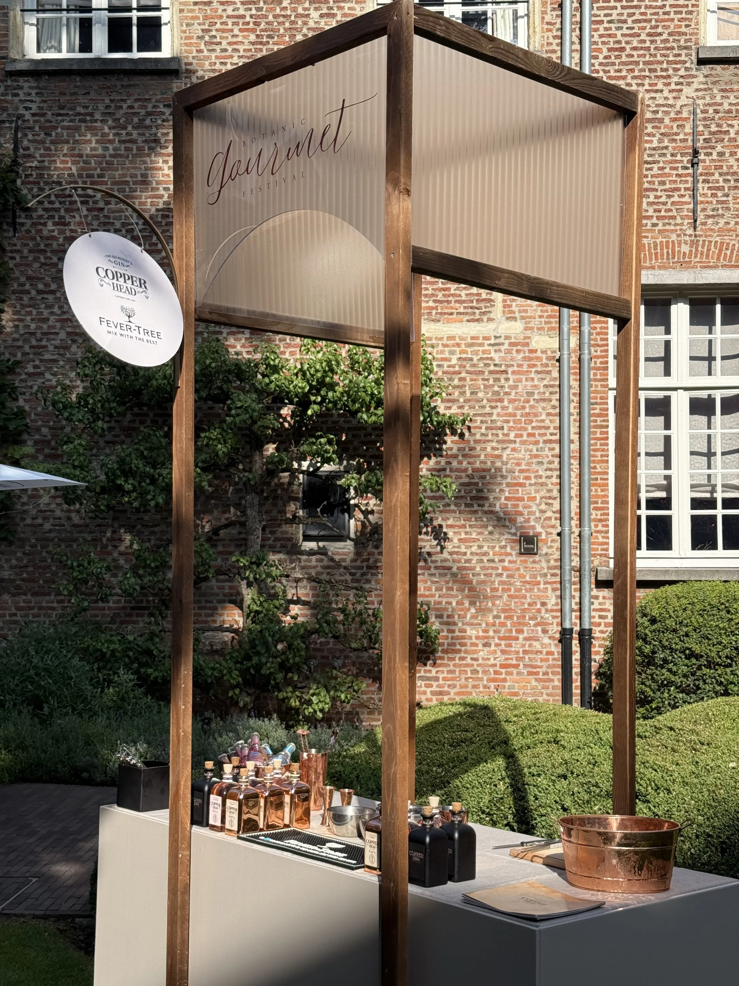 Outdoor display booth at a botanical festival with bottles of gin, copper cups, and glasses on a table, sign reading "Botanic Garden Festival" in the background, brick building and greenery behind.