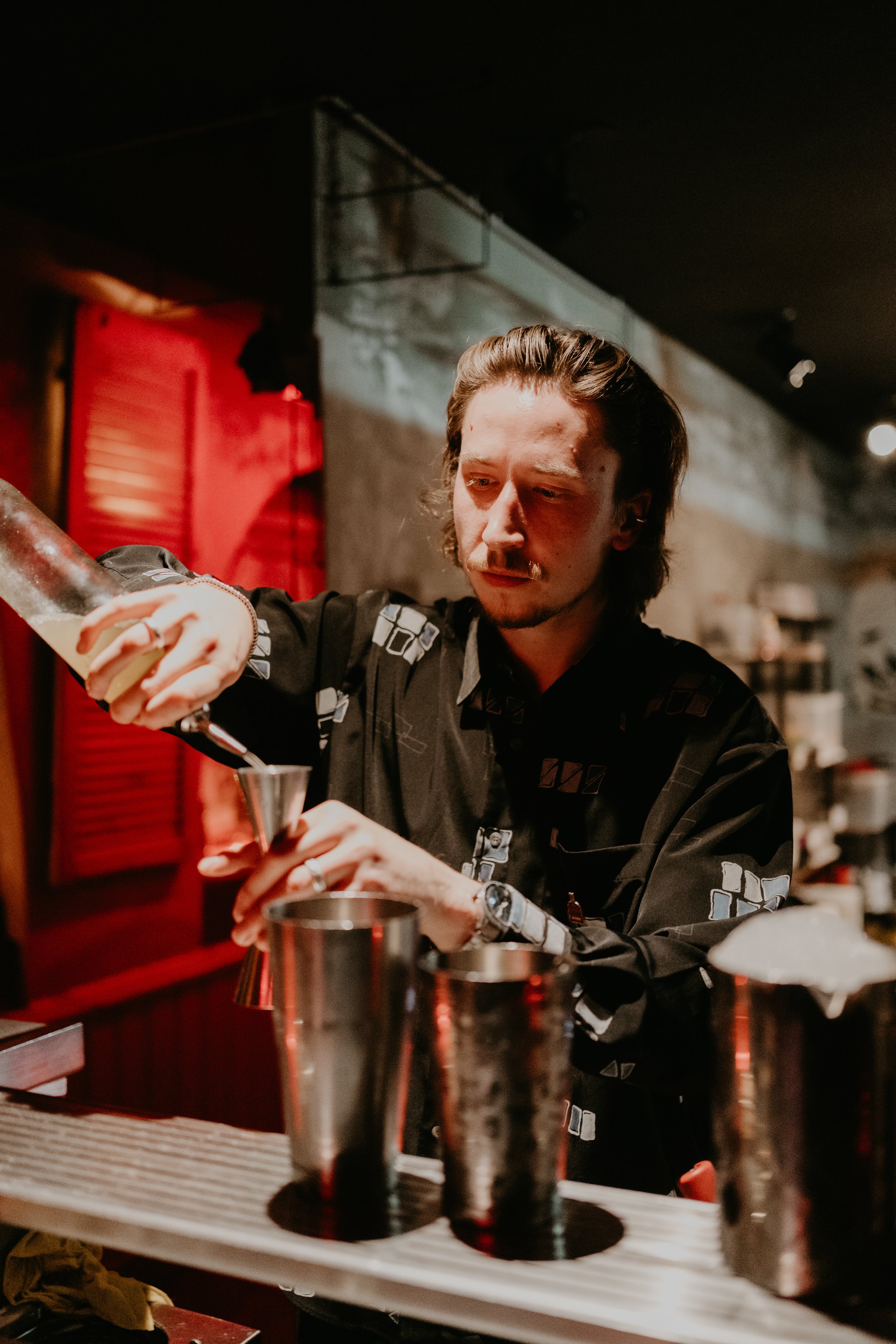 A bartender with long hair in a black patterned shirt pouring a drink through a strainer into a cocktail glass in a dimly lit bar.