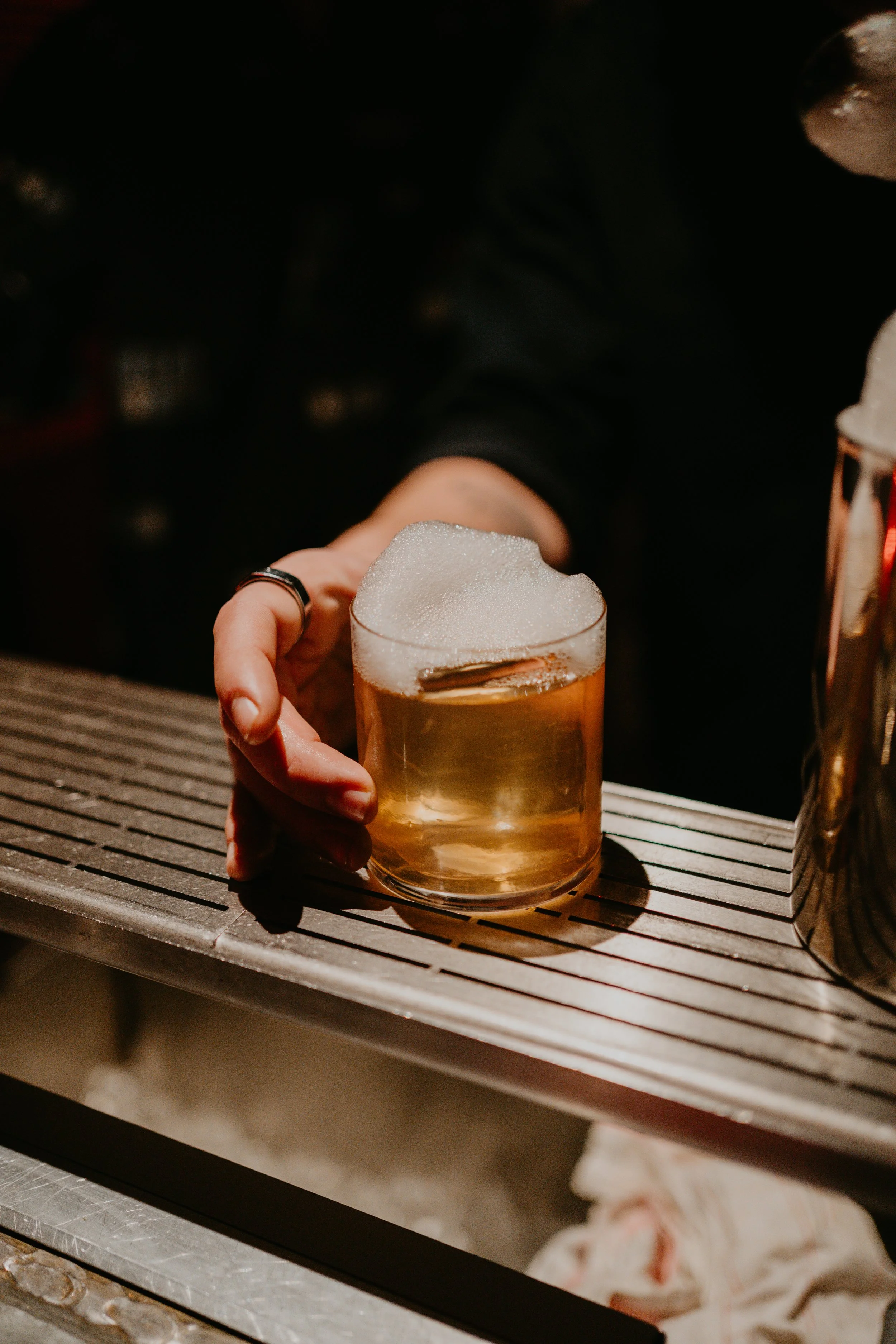 A person holding a glass of beer with foam on top, sitting on a wooden bar counter.