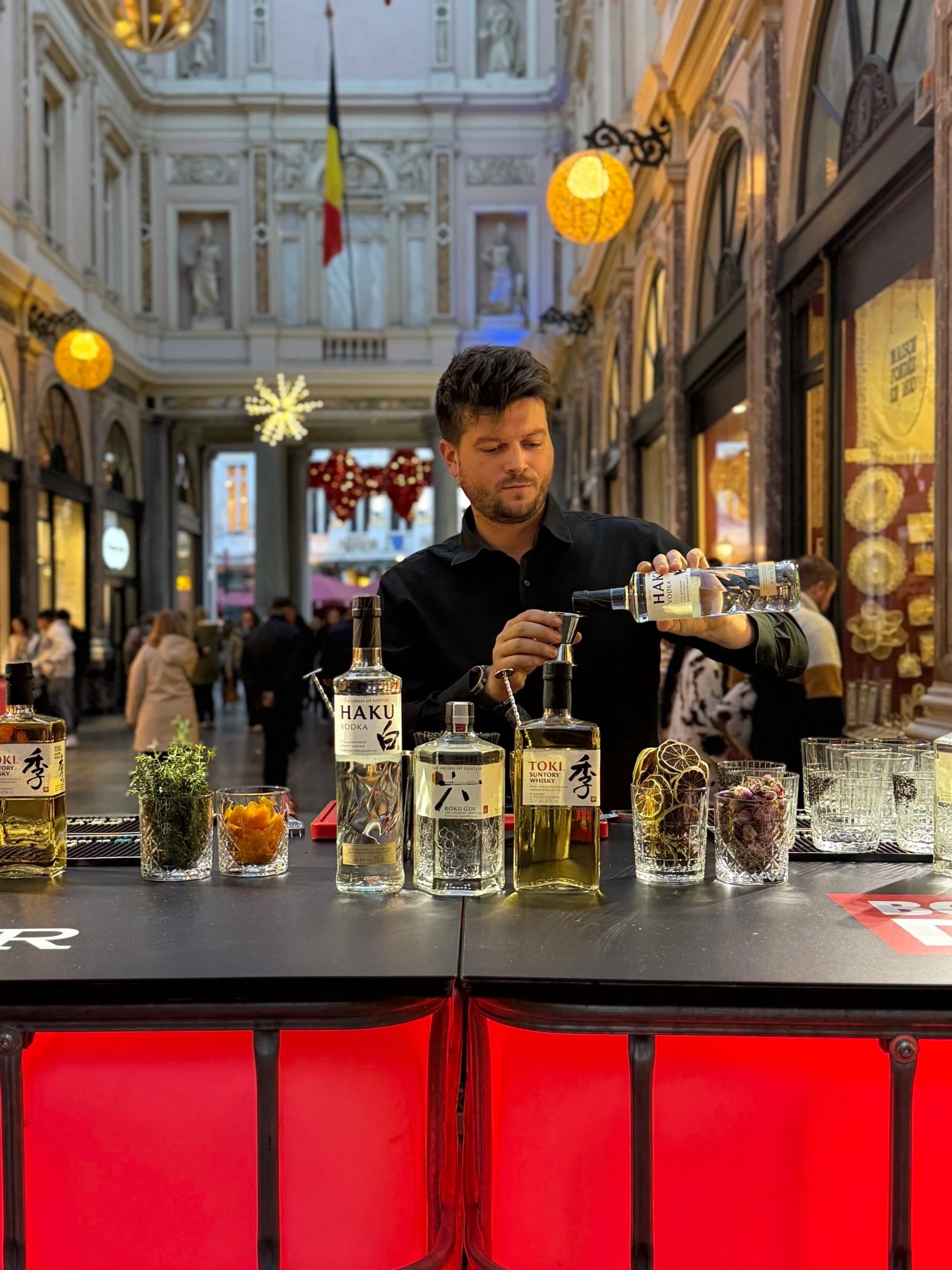A bartender pouring a clear liquor into a glass behind a bar with various bottles and garnishes, inside a shopping arcade decorated with holiday ornaments.