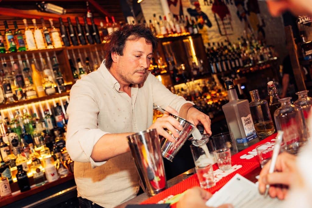 Bartender pouring a drink at a bar with various bottles of alcohol on shelves behind him.