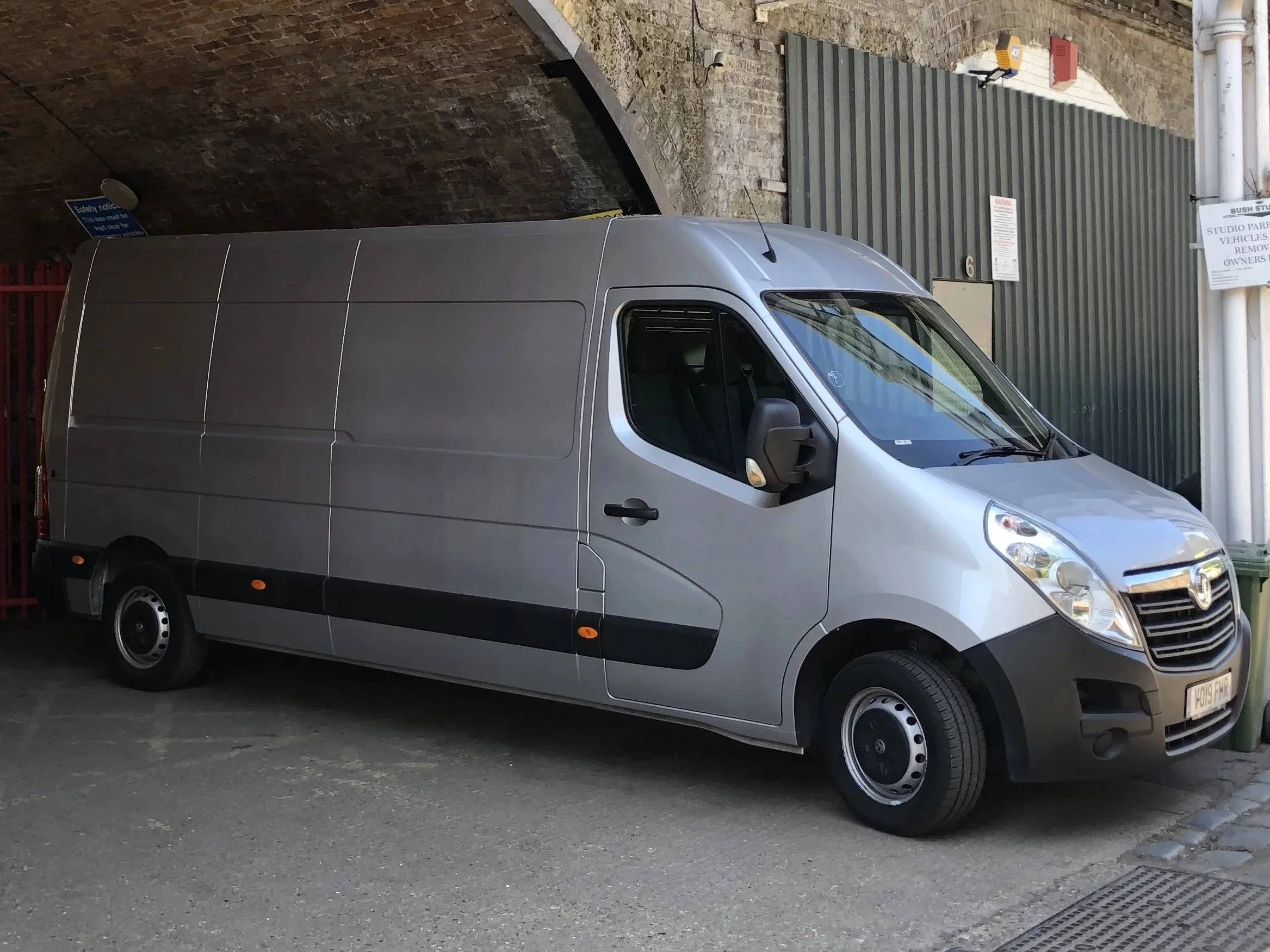 Silver delivery van parked beside a brick wall and a green metal fence under an archway.