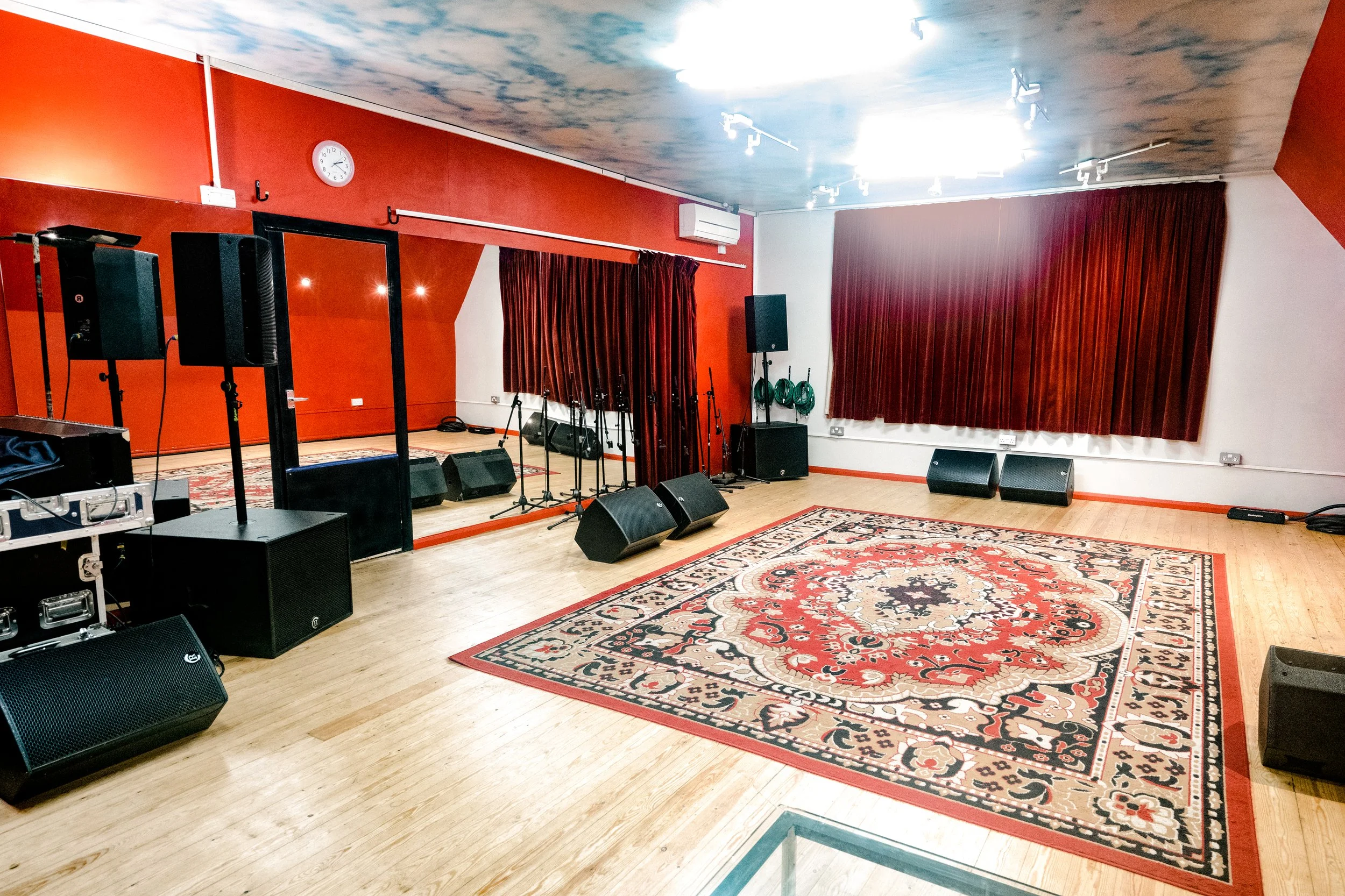 Empty stage with microphones, monitors, and speakers in a room with red walls, curtains, and a decorative rug.