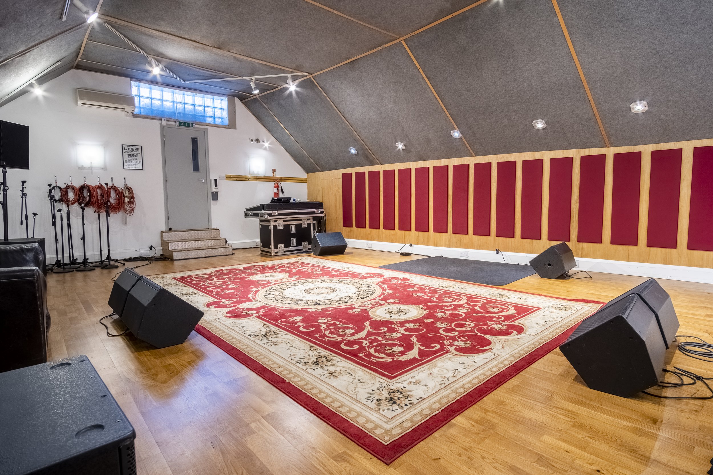 A music rehearsal room featuring a red and beige patterned rug, a sound system with speakers, a black equipment case, wall-mounted acoustic panels, and various microphones and stands, with a gray door and small glass window in the background.