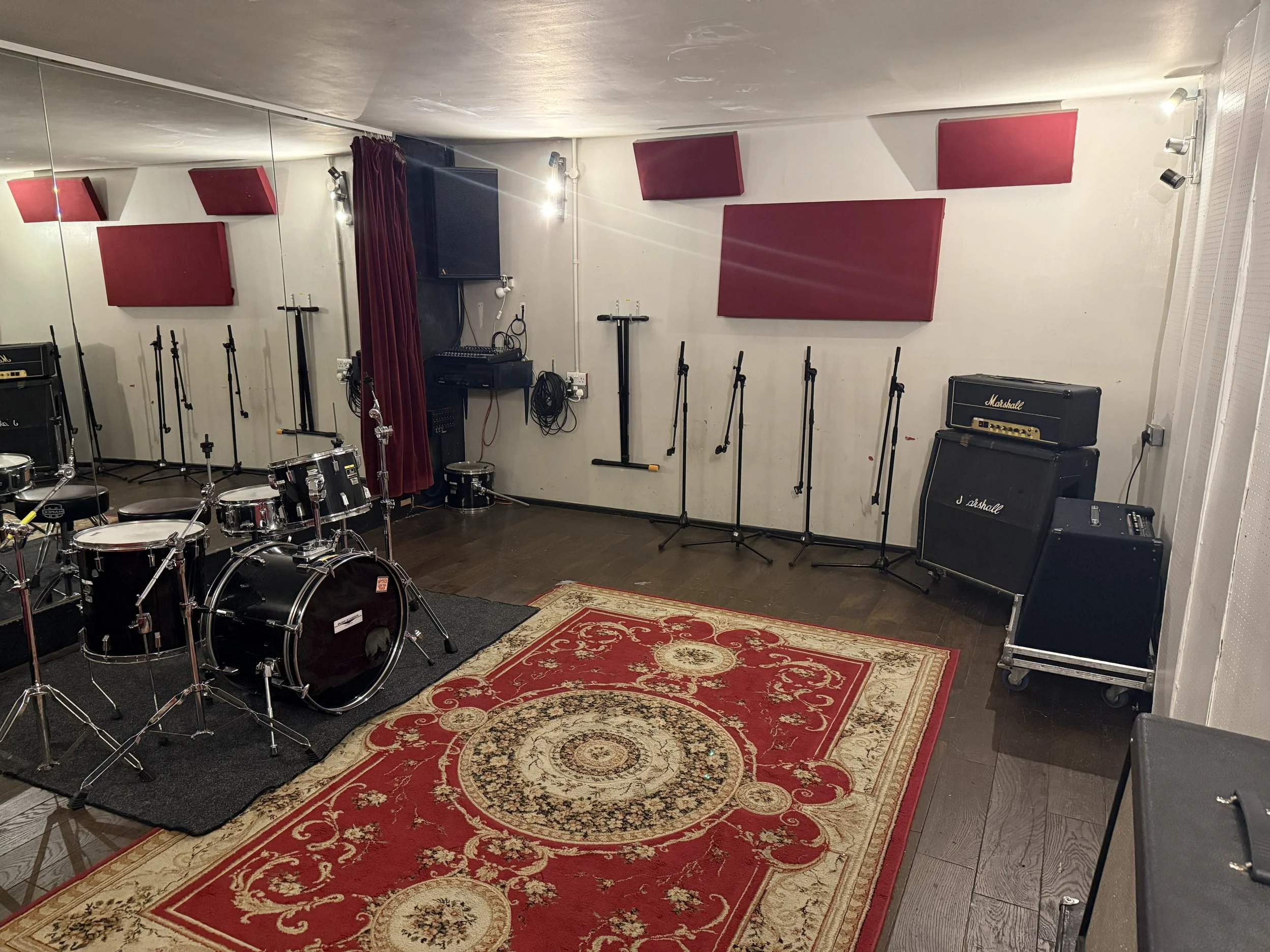 Music practice room with drums, amplifiers, microphones, and acoustic panels, mirror, and rugs.
