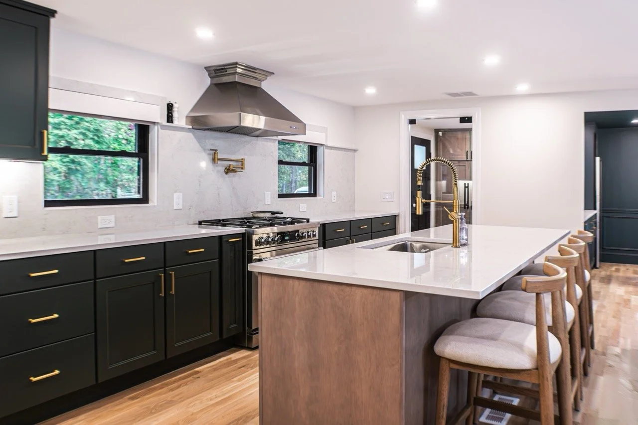 Modern kitchen with black cabinets, white countertops, gold hardware, a central island with a sink and gold faucet, bar stools, stainless steel appliances, and windows overlooking green trees.