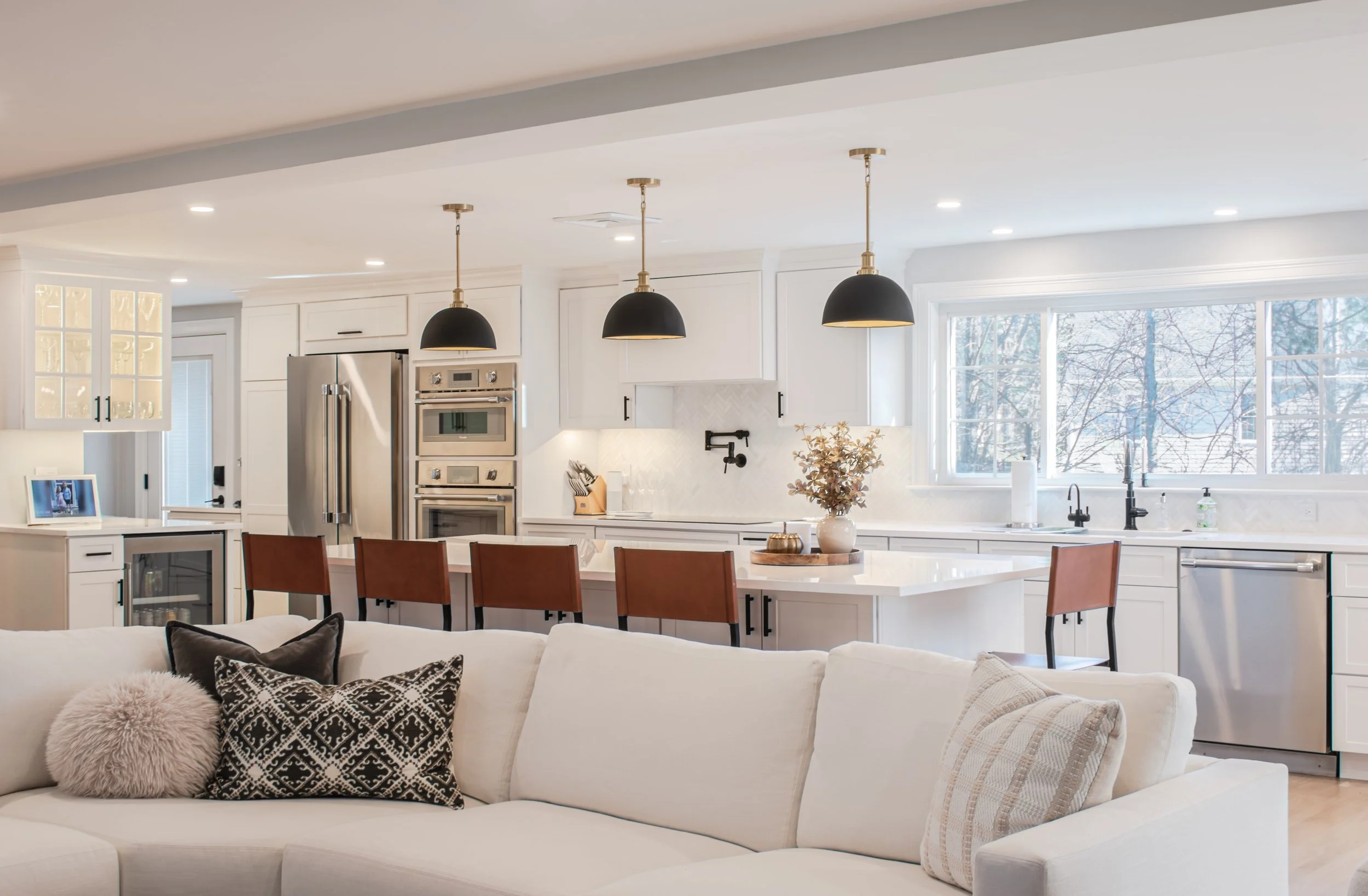 Open concept kitchen with white cabinets, stainless steel refrigerator and double oven, black pendant lights, large window, and a white sofa with decorative pillows in the foreground.
