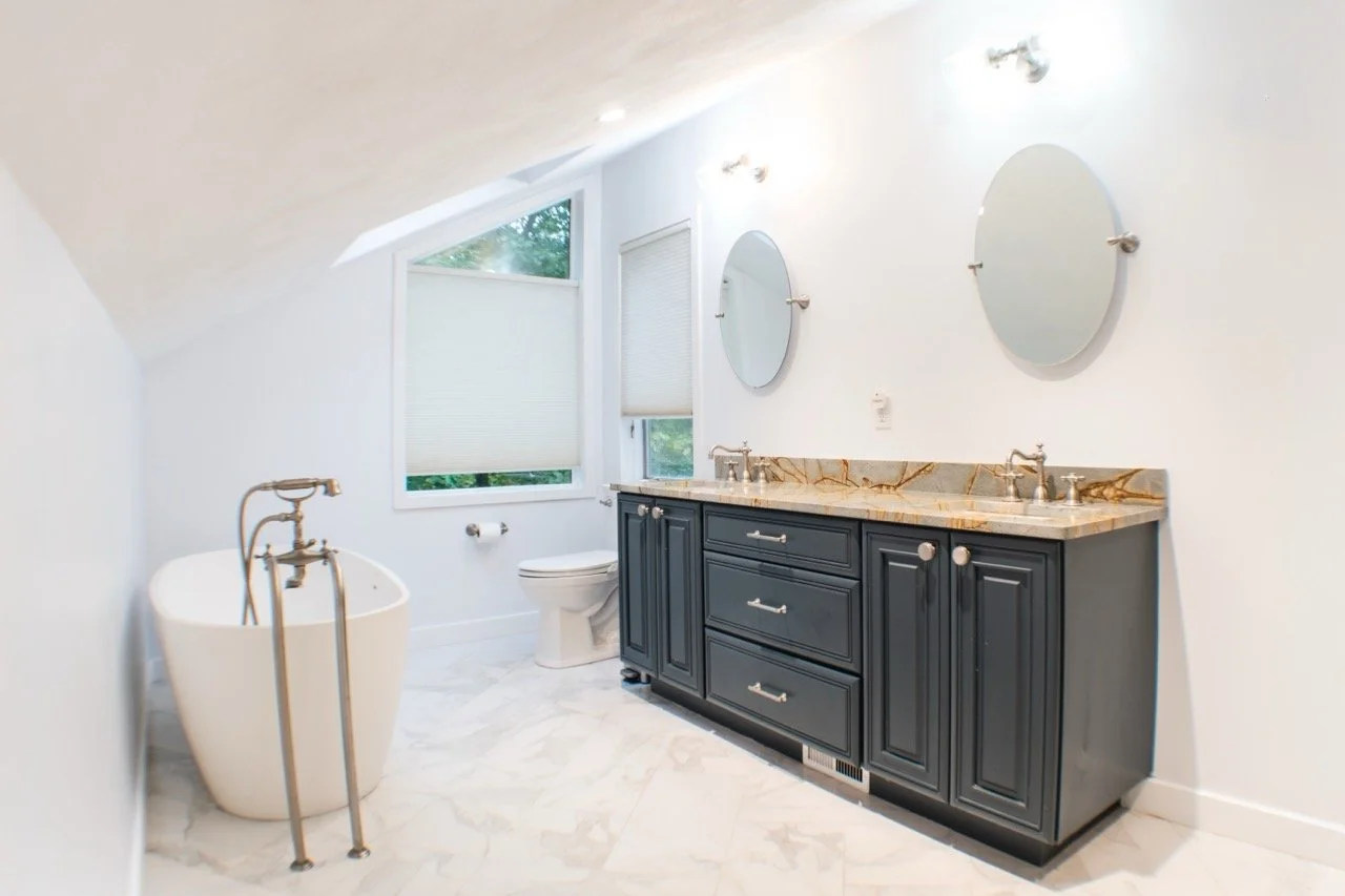 Modern bathroom with white walls, marble floor, a black vanity with two sinks, oval mirrors, a toilet, large window with blinds, and a freestanding bathtub.