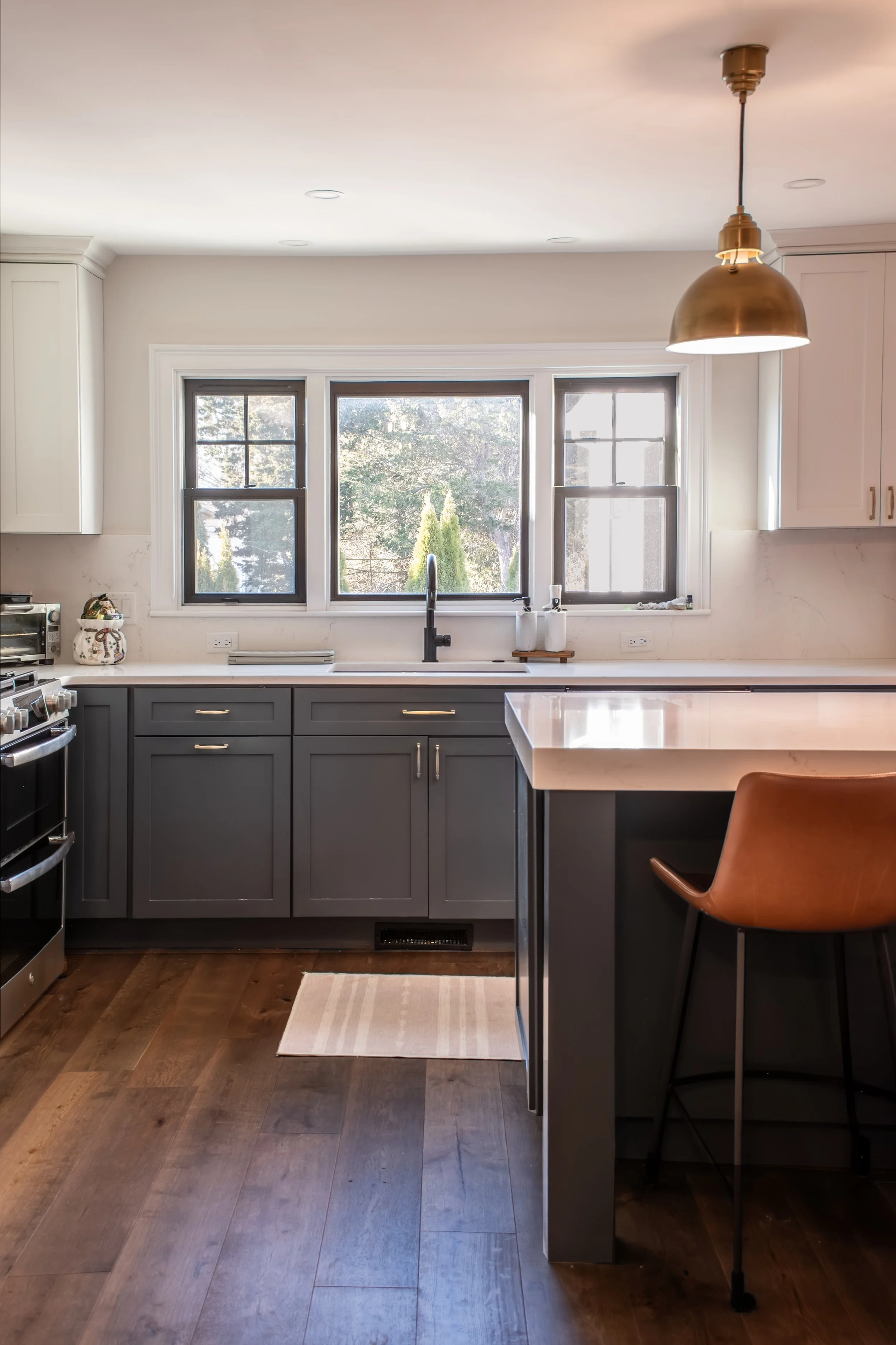 Modern kitchen with gray cabinets, white countertops, a window over the sink, a gold pendant light, and a brown barstool at the counter.