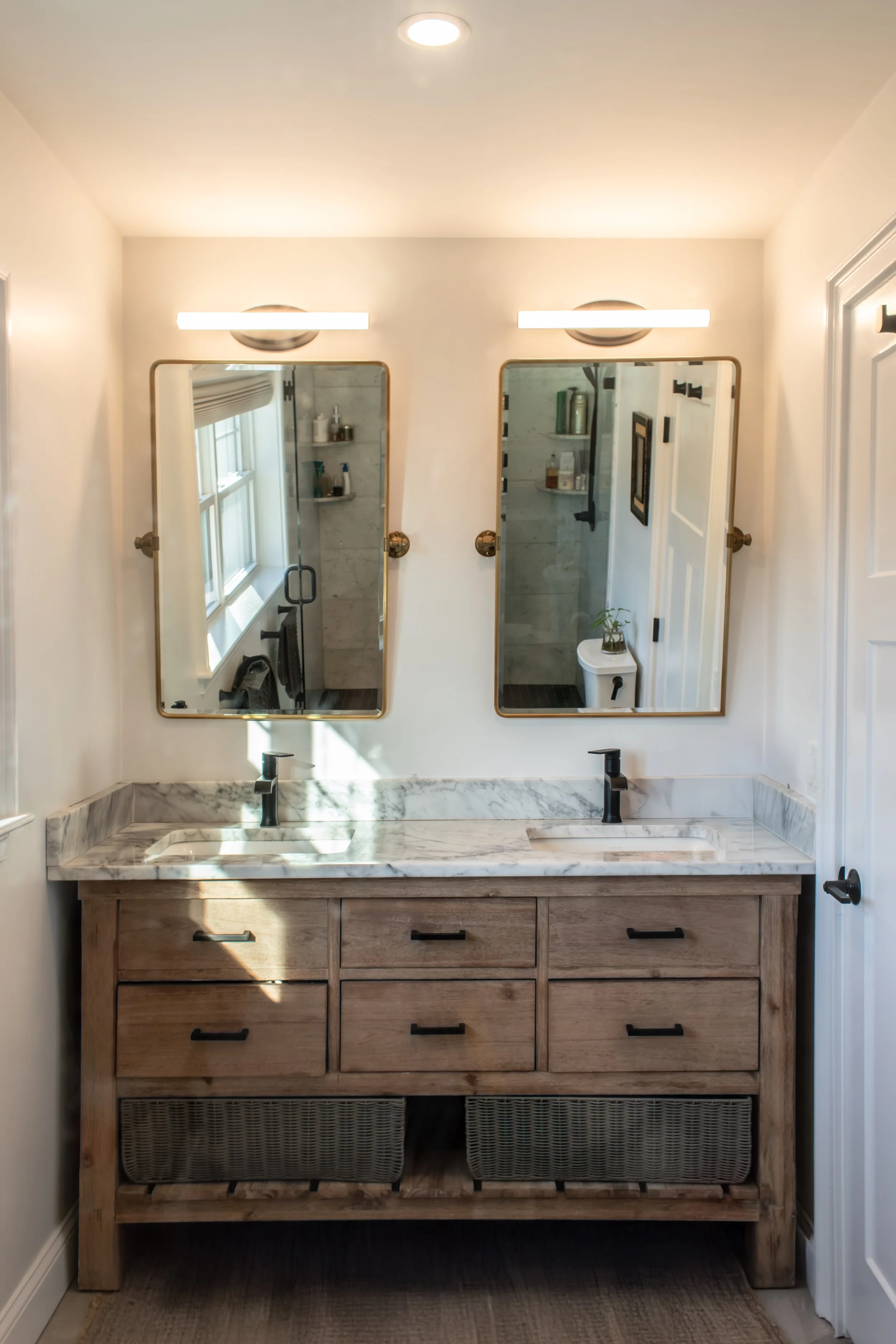 Bathroom with a wooden vanity, marble countertop, two black sinks, and two mirrors with gold frames, illuminated by two horizontal light fixtures.