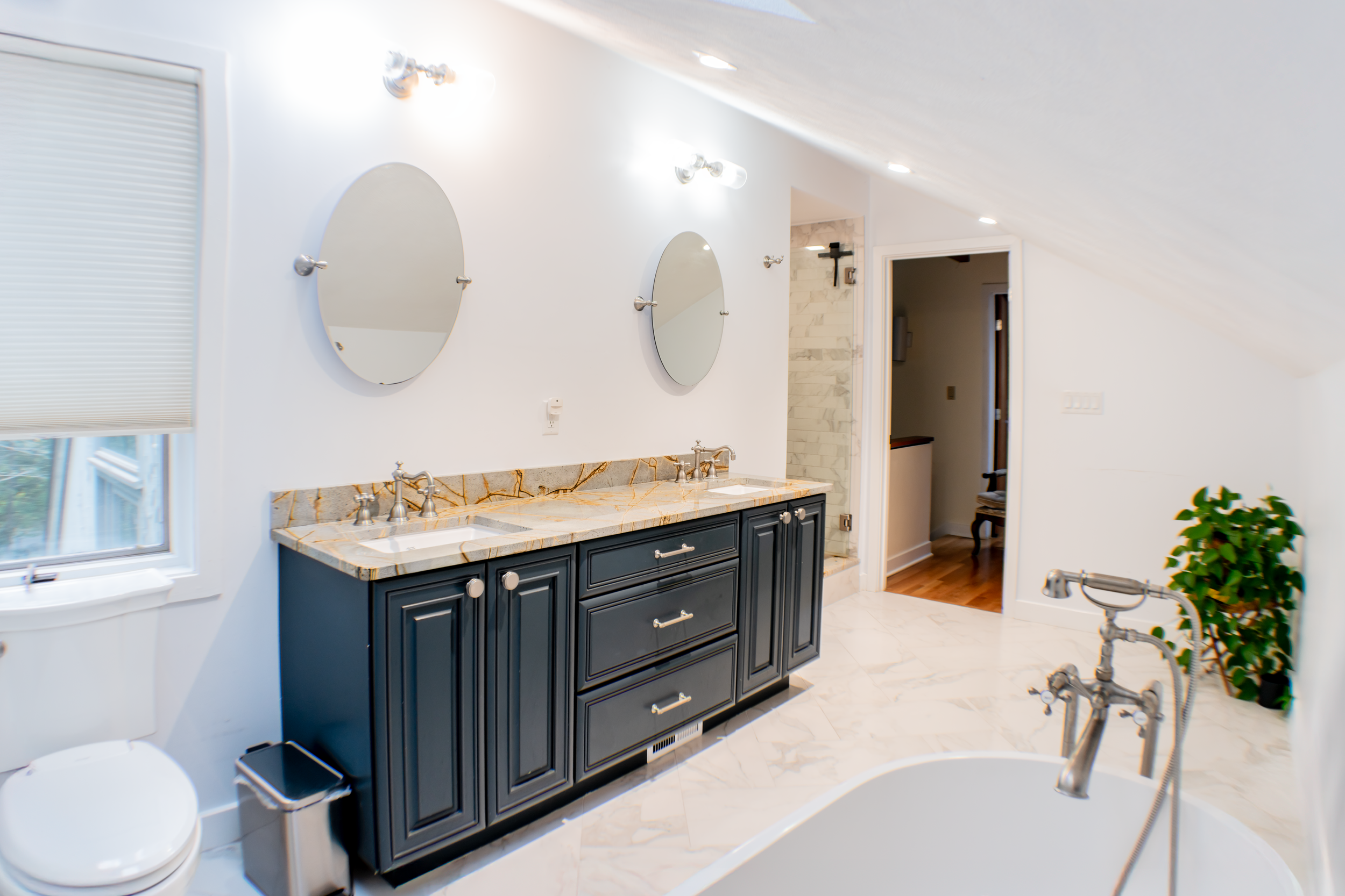 Modern bathroom with a white freestanding bathtub, a toilet, and a dark gray vanity with two sinks and two round mirrors. White walls, marble floor, and a window with blinds.
