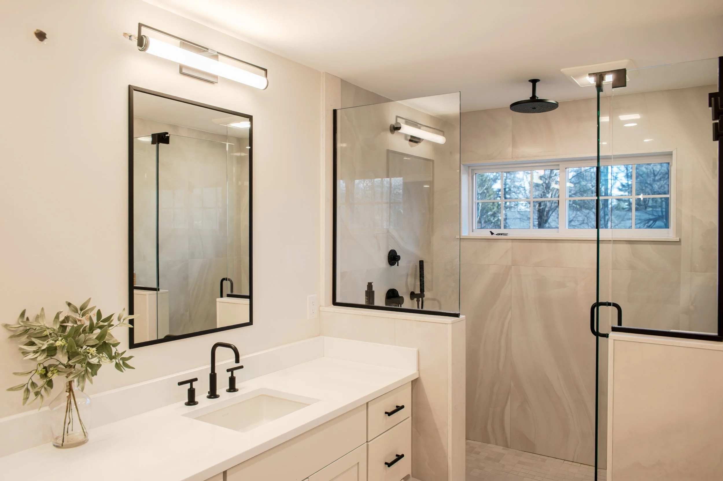 Modern bathroom with white vanity, black fixtures, large mirror, and glass-enclosed shower with beige marble walls and a window.