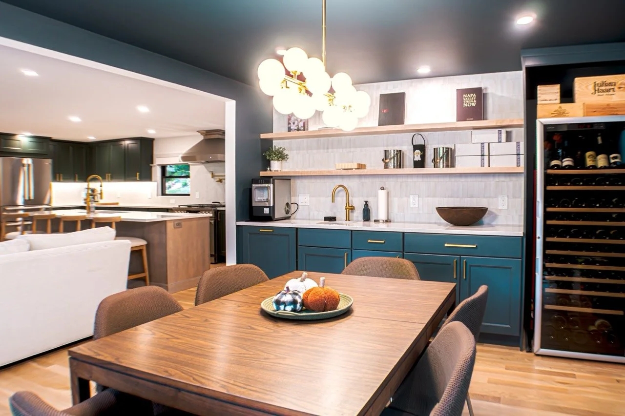 Modern kitchen and dining area with a wooden table, beige chairs, and teal cabinets. Open shelving with decor, a wine fridge, and a chandelier overhead.