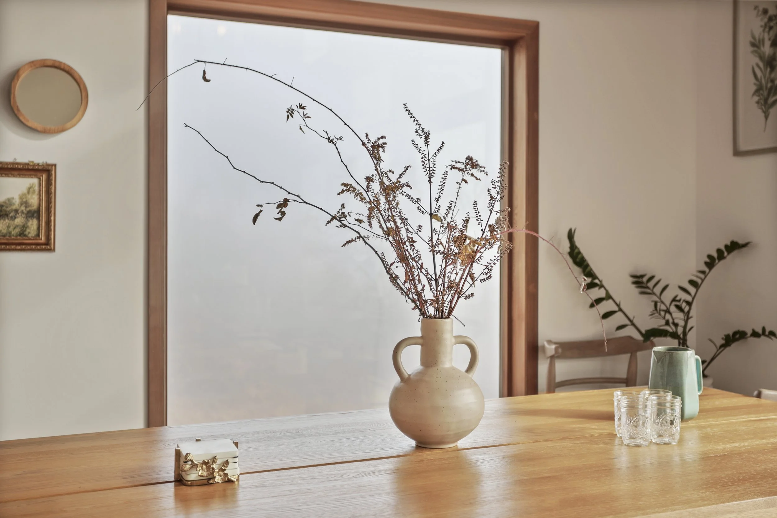A dining table with a beige ceramic vase containing dried branches and foliage, a small box with a bow, three glass cups, a green pitcher with a green plant, and a wooden chair in a room with a large window and framed artwork on the walls.