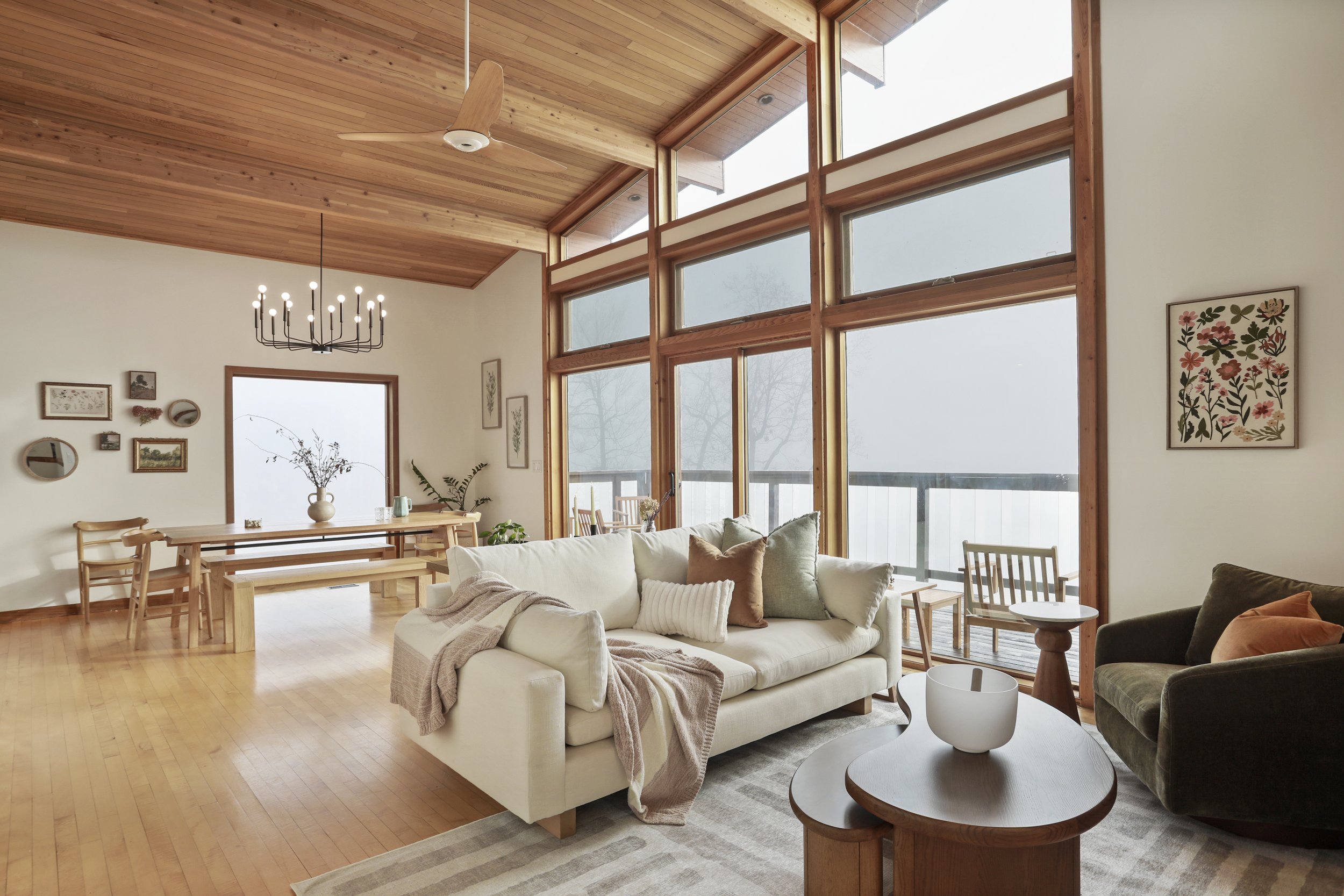 Living room with large windows, white sofa, dark armchair, wooden ceiling, and decor including framed artwork, plants, and a chandelier.