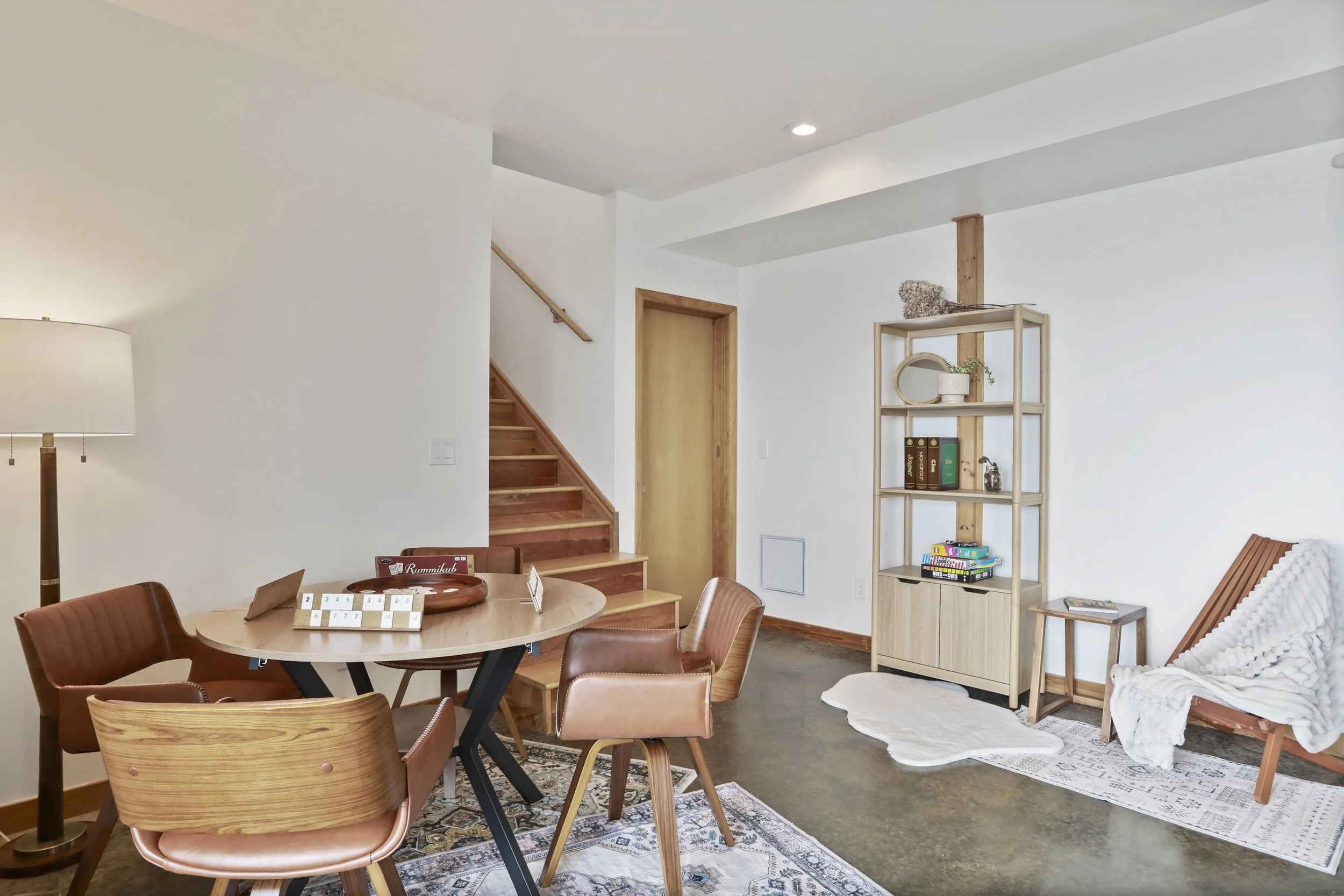 Living room with a round wooden table, four brown chairs, a wooden staircase, a bookshelf with decorative items, a floor lamp, and a cushioned chair with a white throw blanket.