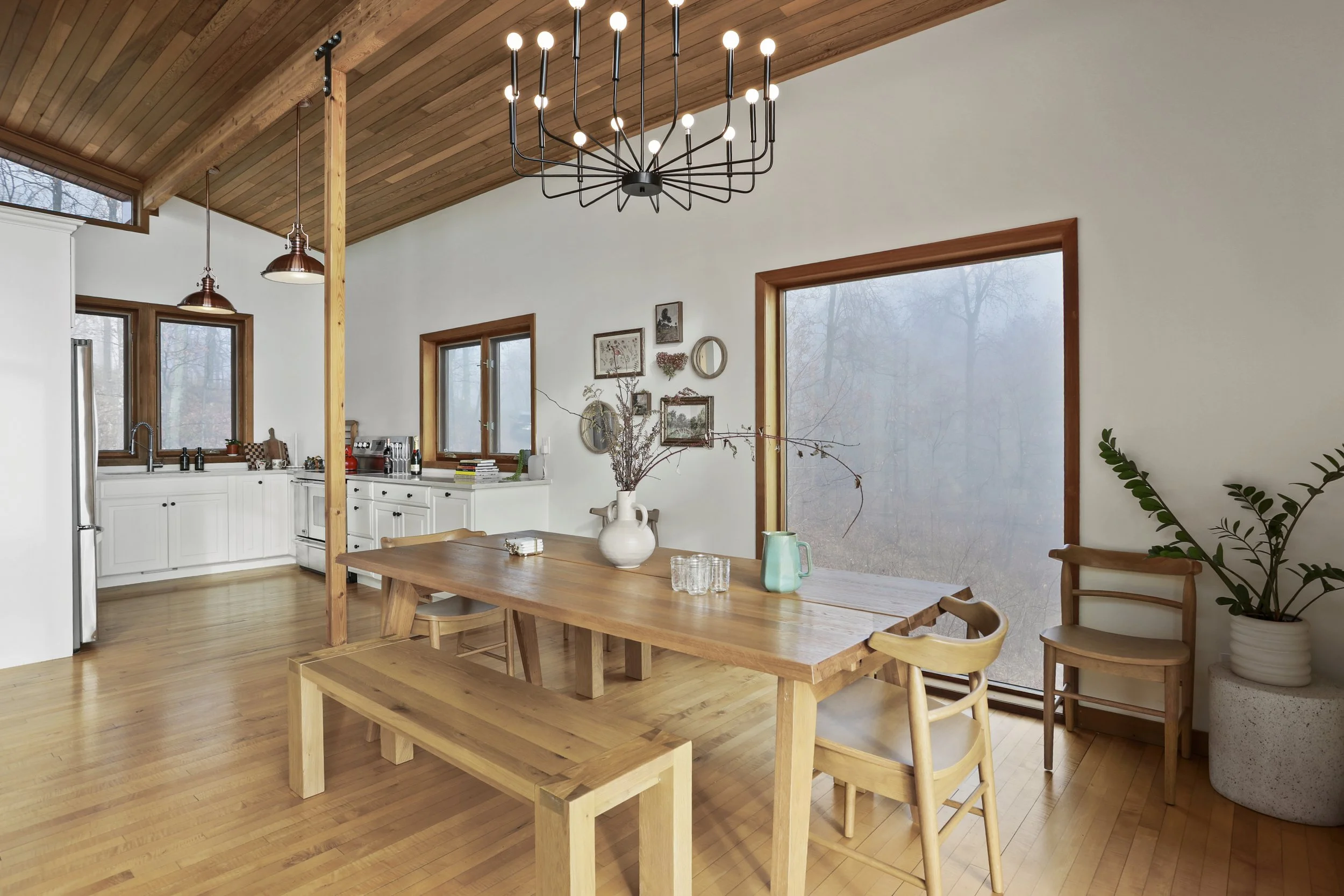 A modern dining room with wooden furniture, a large wooden table with seating, and a view of a kitchen with white cabinets. There's a large window and glass door showing a foggy outdoor scene with trees. The room has wooden flooring, a wooden ceiling