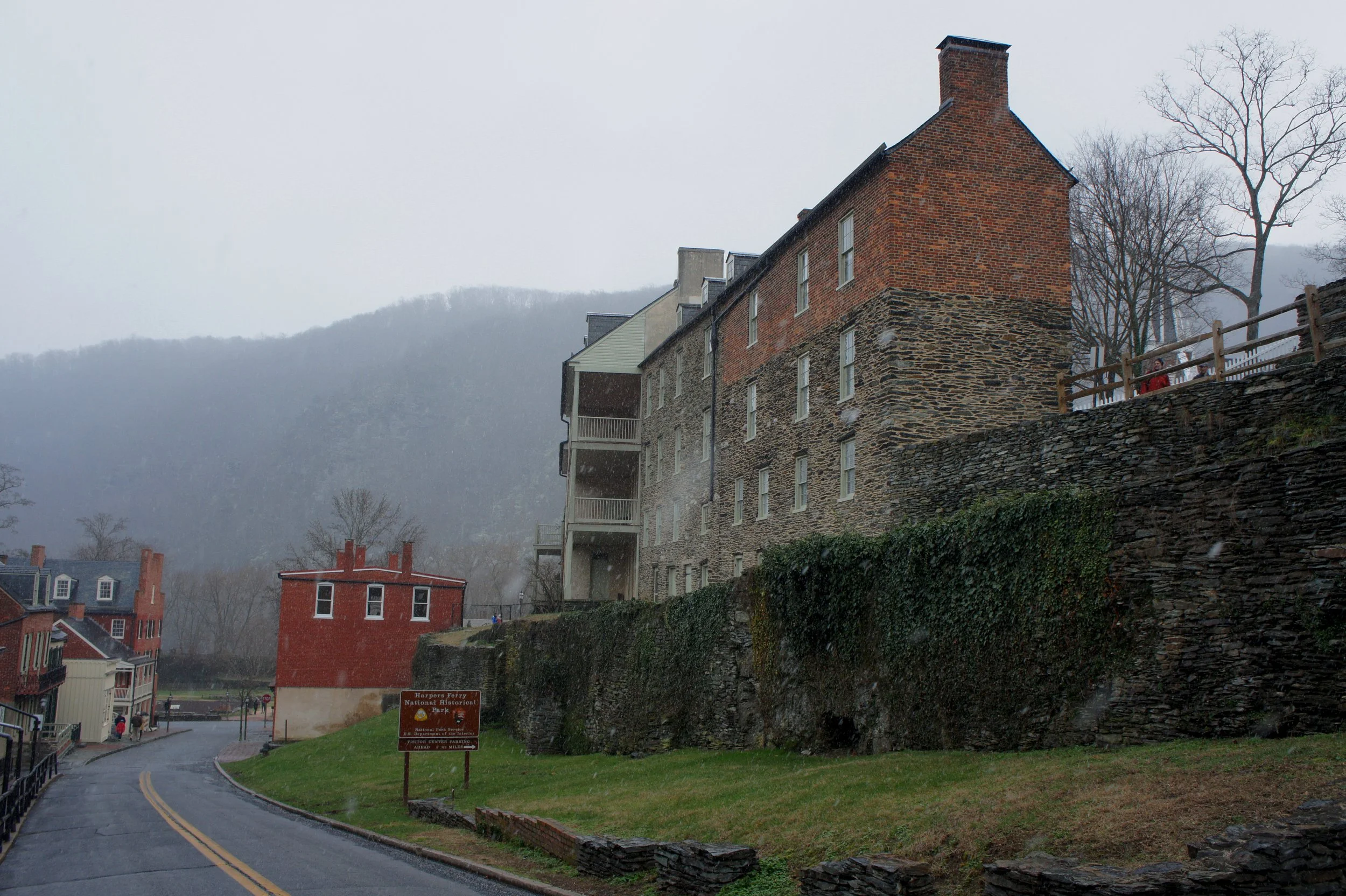 Historic brick and stone buildings along a curved street with a sign indicating Harper Ferry National Historical Park, misty hills in the background on a rainy day.