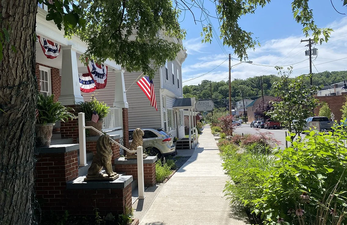 Sidewalk view in a small town with American flags, lion statues, and a building with a porch decorated with patriotic bunting. Trees and greenery line the street, with several parked cars and a sign reading 'Atasia Spa' visible in the background.