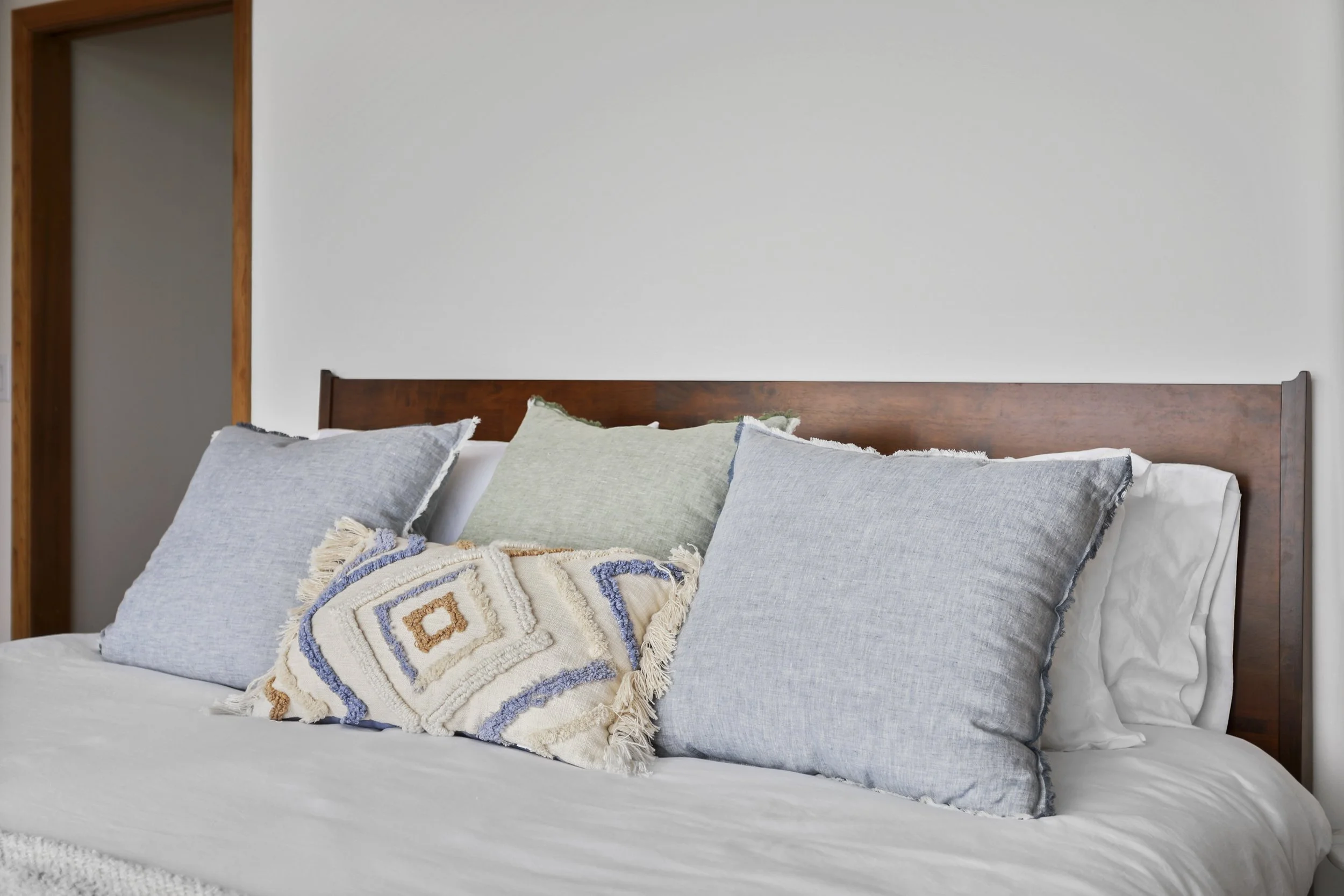 A bed with six pillows, including two large blue pillows, one green pillow, a white pillow behind the green one, and a decorative pillow with geometric patterns. The headboard is wooden, and the wall behind is plain and light-colored.