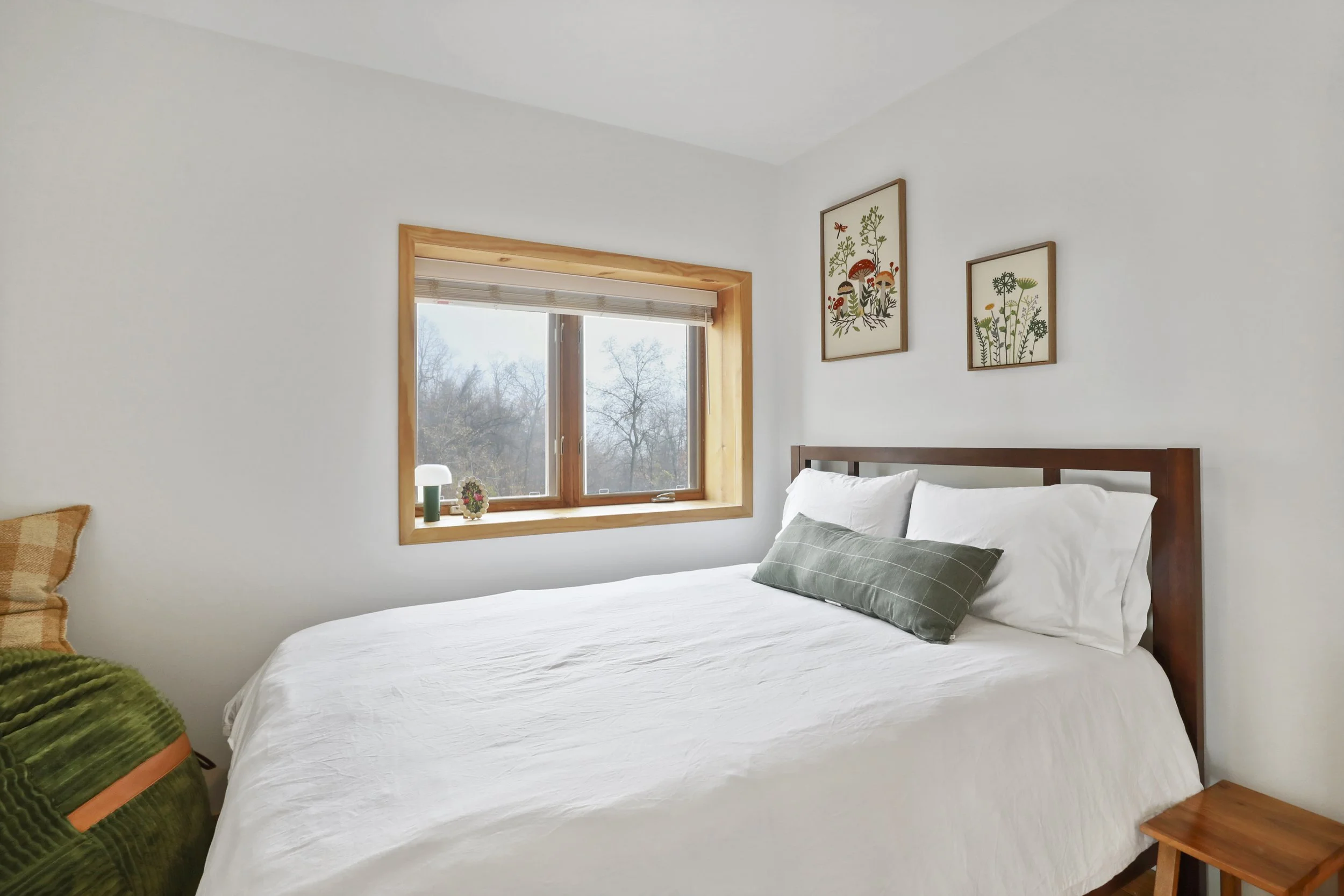 A cozy bedroom with a bed, white bedding, green decorative pillow, wooden headboard, and artwork of plants and mushrooms on the wall, next to a window with a view of leafless trees.