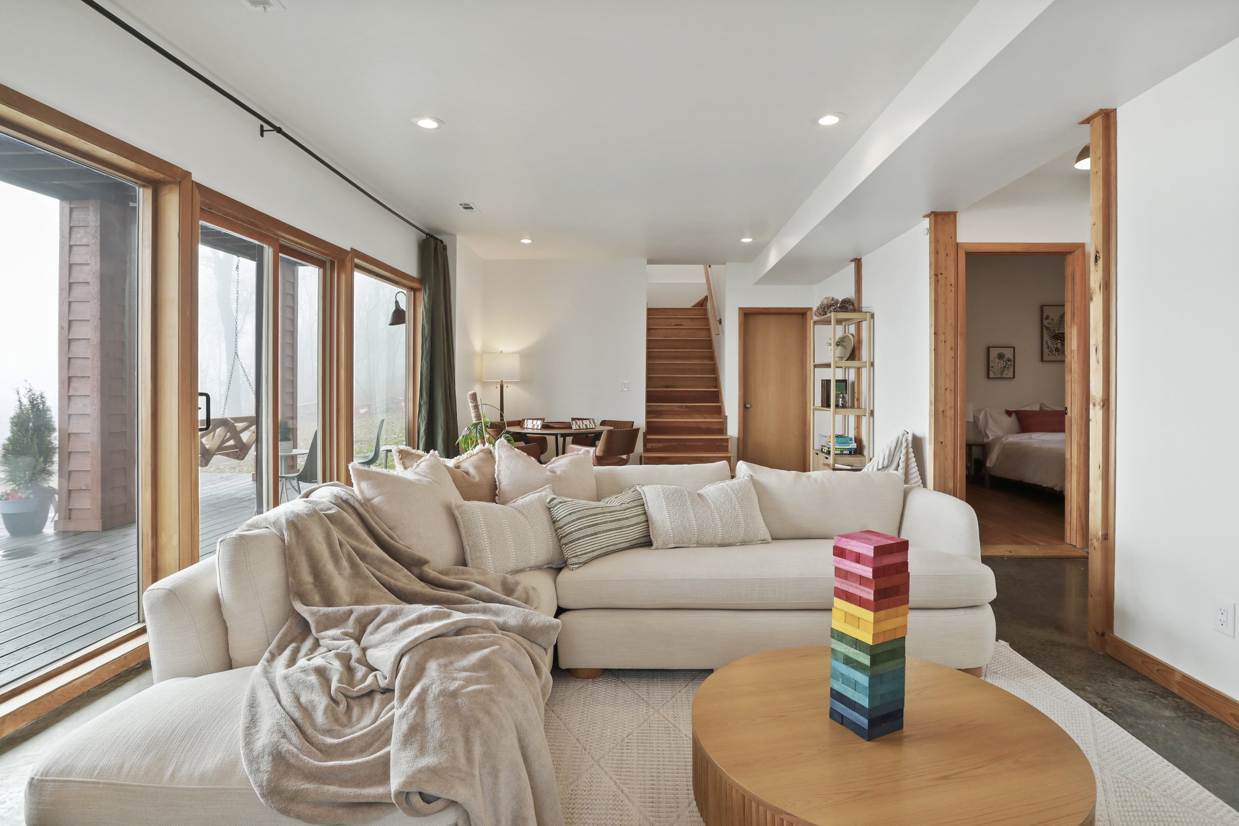 Living room with a beige sectional sofa, beige pillows, a blanket, a wooden coffee table with a colorful tower toy, large sliding glass doors leading to an outdoor porch, a dining area with a round table and chairs, wooden stairs, a bookshelf, and a 