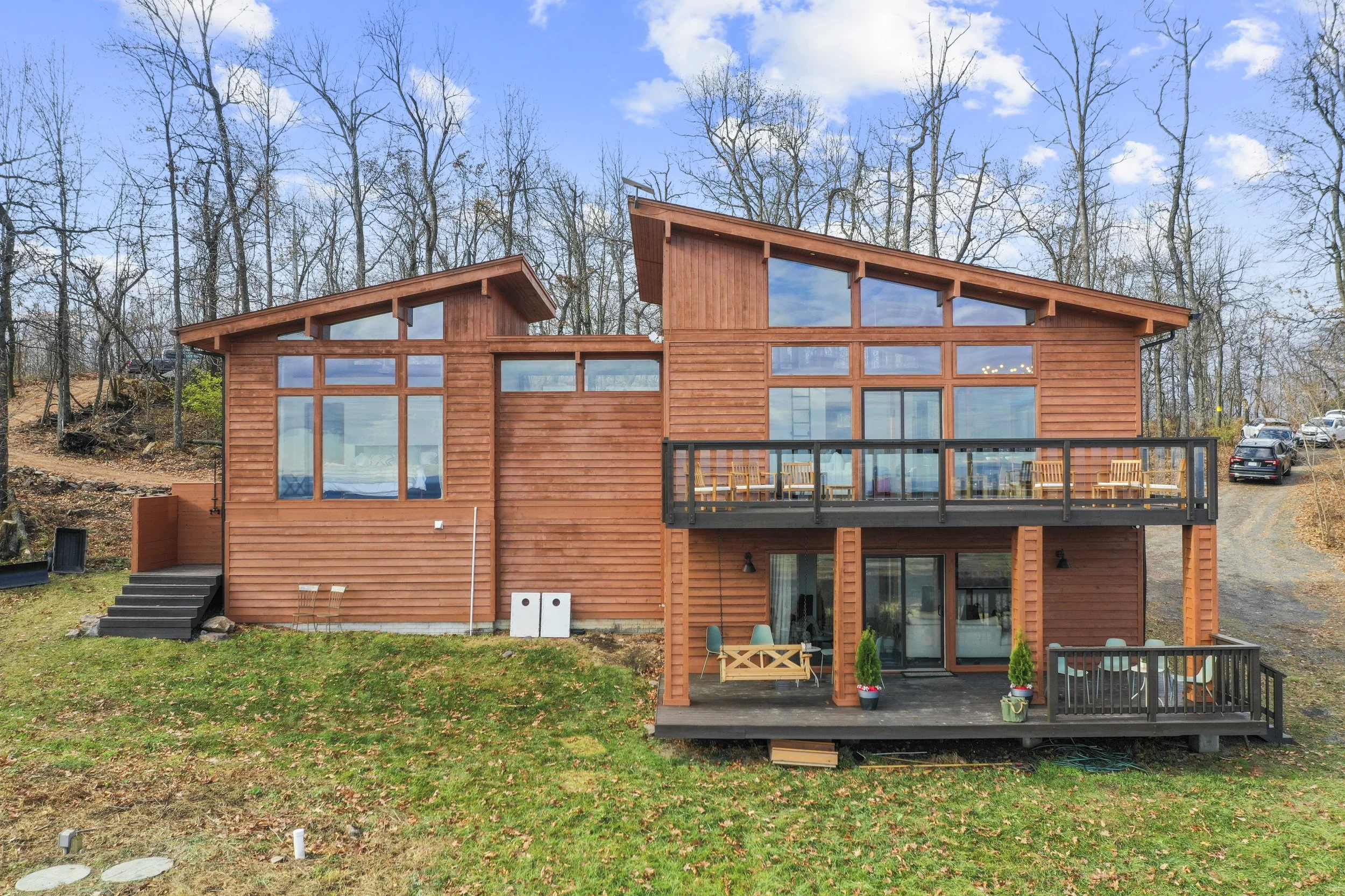 Large modern two-story wooden house with large windows, multiple decks with outdoor furniture, set in a wooded area with leafless trees, under a blue sky with some clouds.
