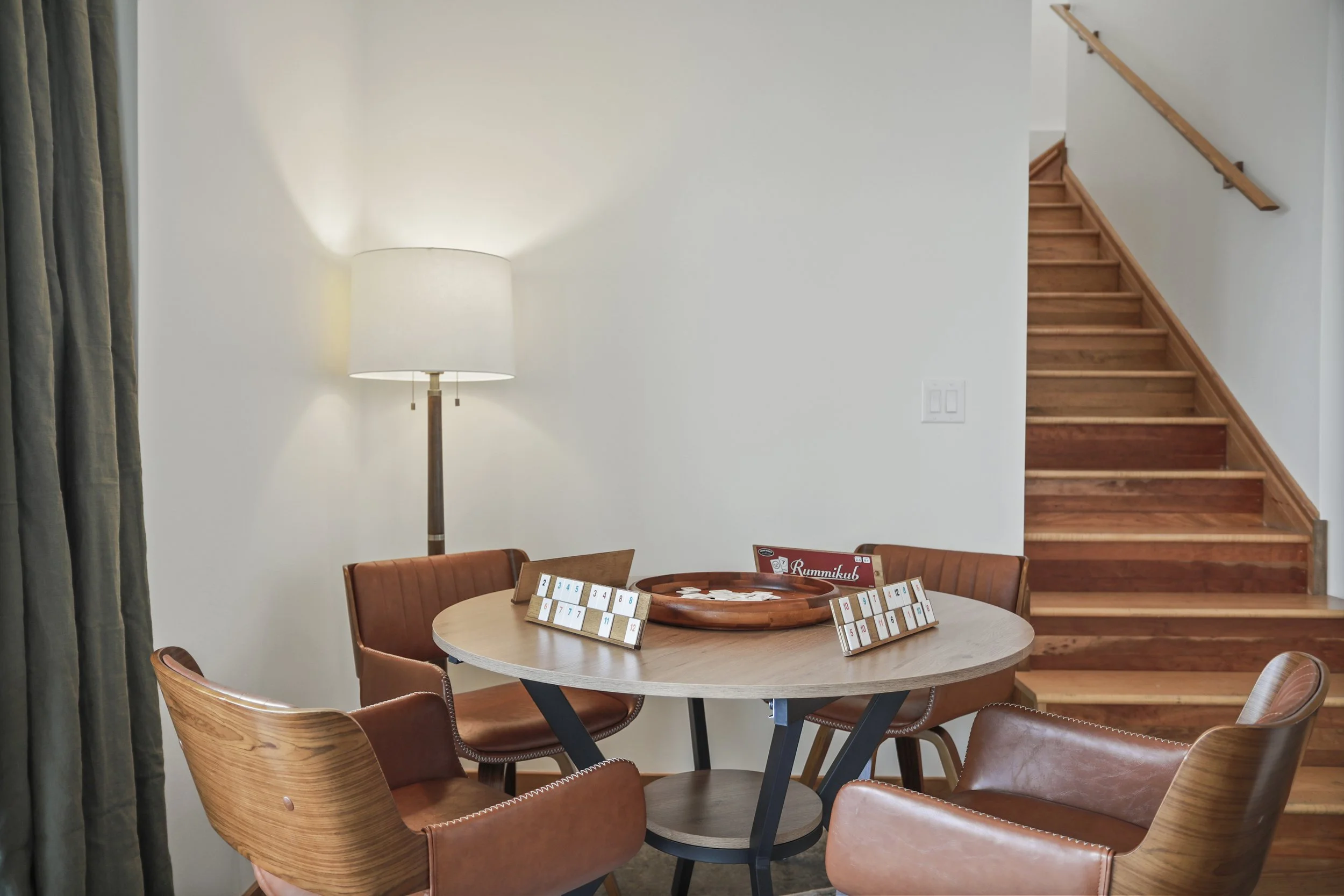 A round wooden table surrounded by four brown leather chairs with a game of Rummikub on top, a tall floor lamp next to the table, and a staircase in the background.