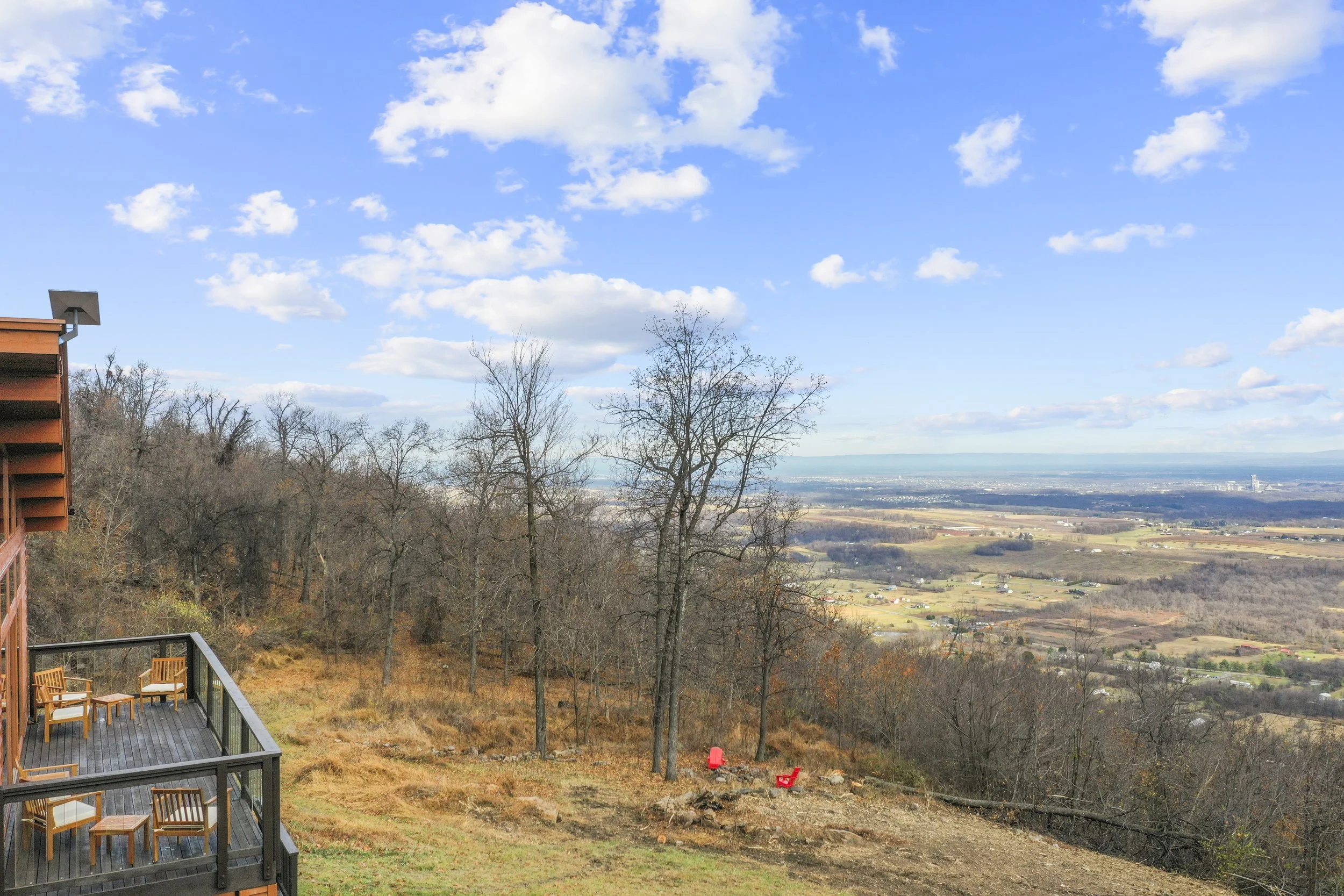 View from a hillside with a wooden deck, chairs, bare trees, and a valley with fields in the distance under a partly cloudy blue sky.