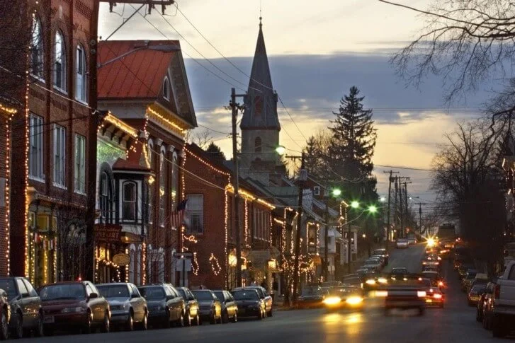 A street scene at dusk in a small town with Christmas decorations, parked cars, a church steeple, and some cars driving with headlights on.