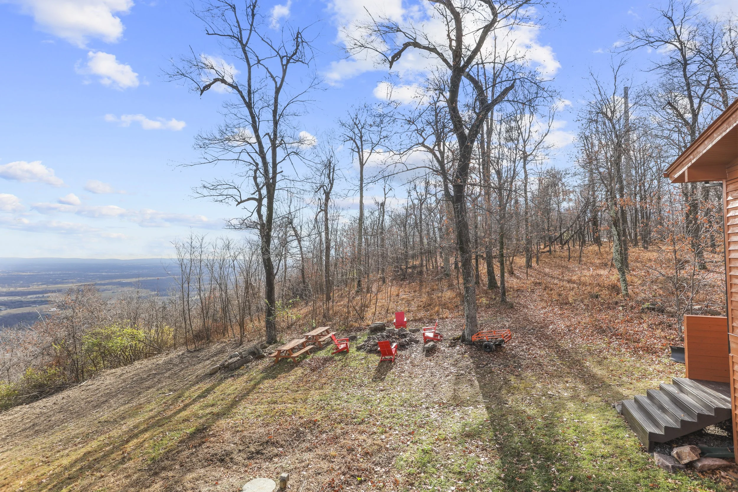 A hillside backyard with leafless trees, wooden benches, red Adirondack chairs arranged around a campfire, a small orange wagon, and a house with stairs on the right, overlooking a distant landscape with a blue sky and clouds.