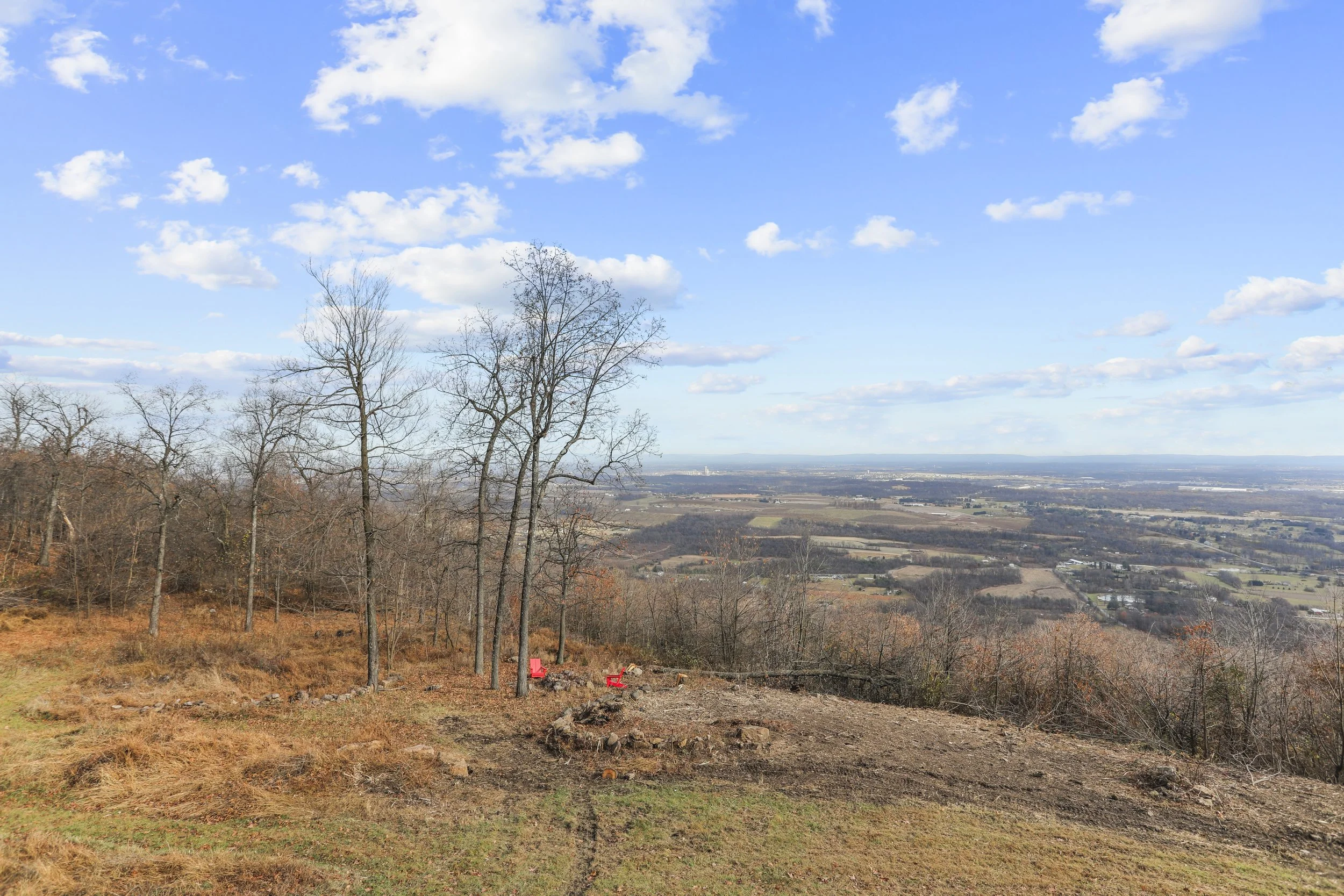 Scenic view from a hilltop with a few leafless trees, orange chairs on cleared earth, and a distant city skyline under a partly cloudy blue sky.