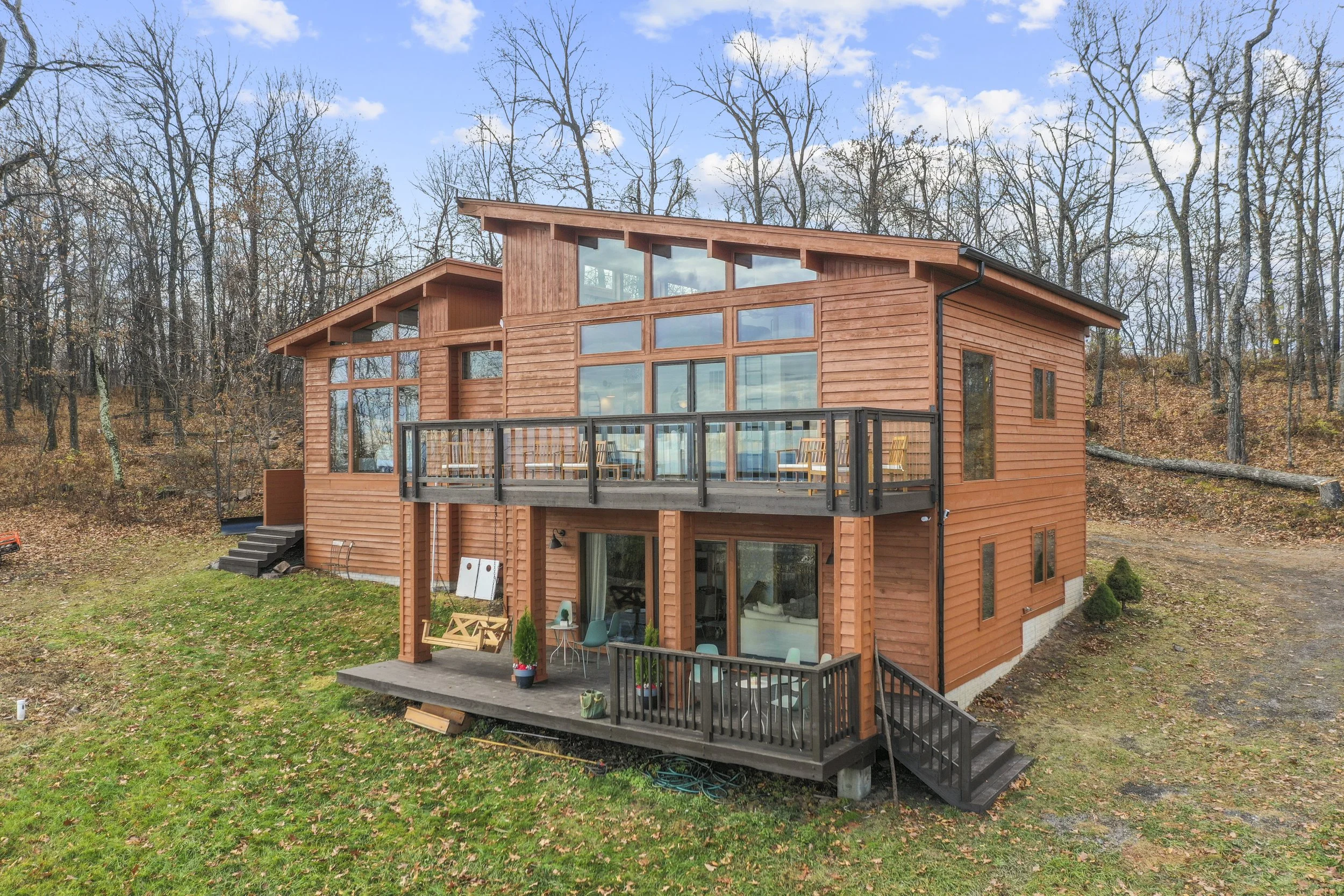 A modern three-story wooden house with large windows and balconies, situated in a wooded area with trees and fall foliage, under a partly cloudy sky.