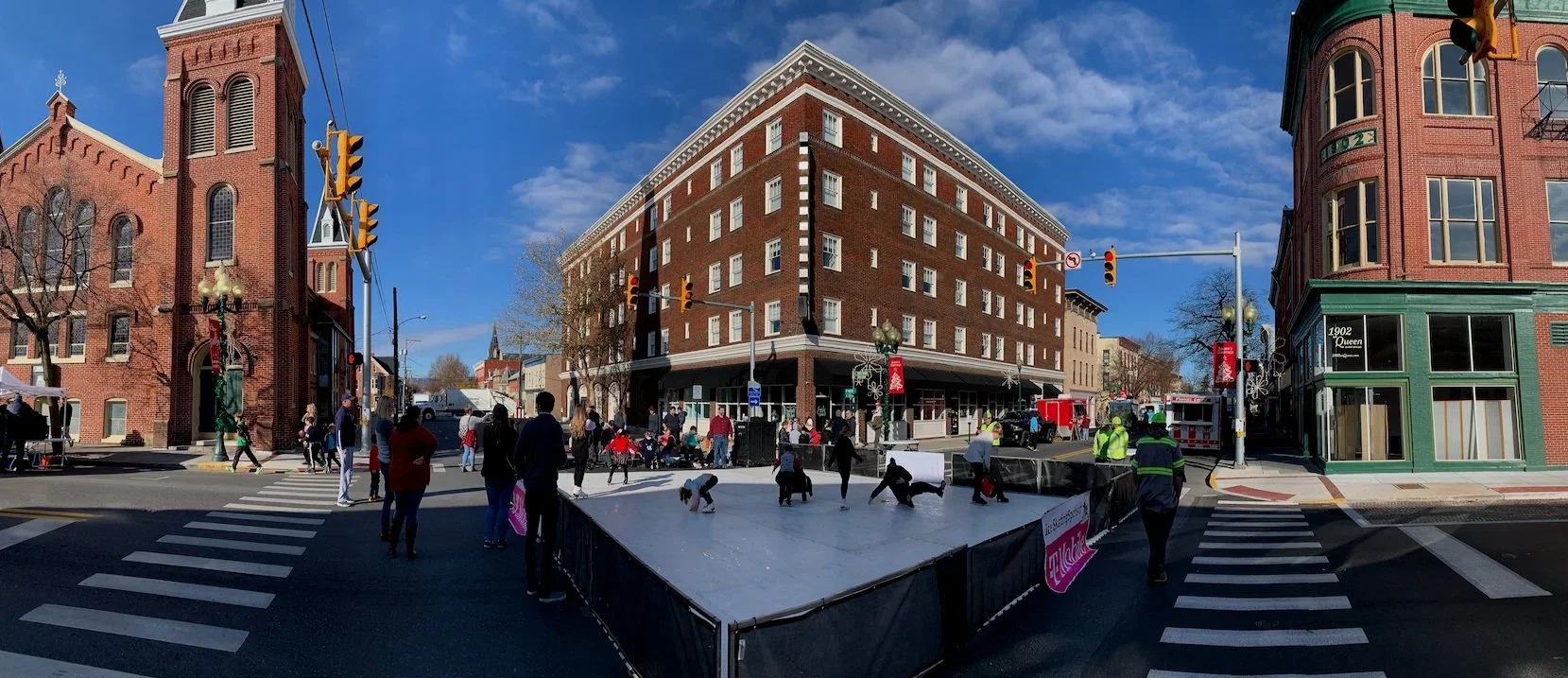 People ice skating outdoors on a small rink at a street intersection, surrounded by brick buildings, with blue sky and some clouds overhead.