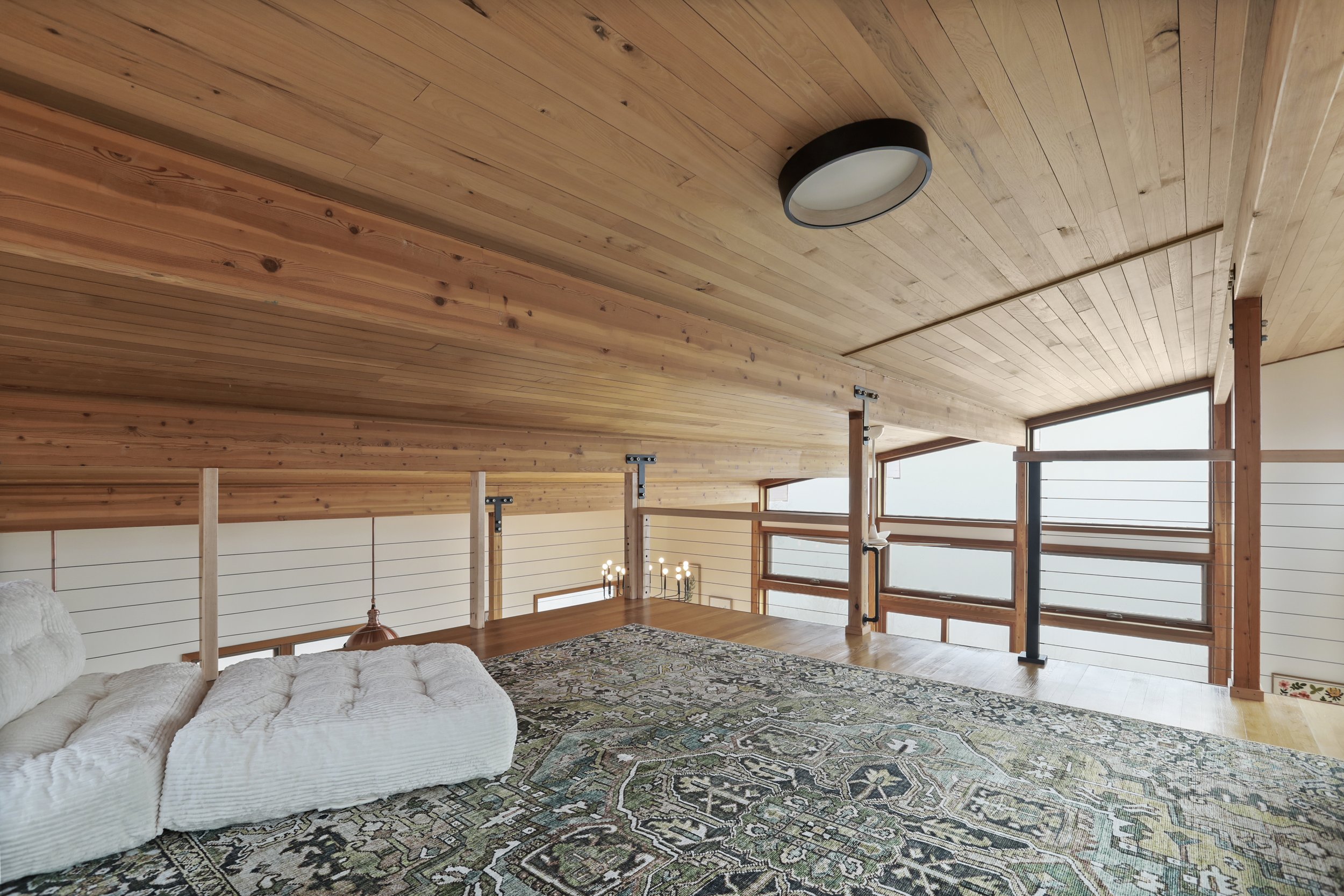 A cozy interior space with wood-paneled ceilings and walls, large windows, a plush white sofa, and a patterned area rug.