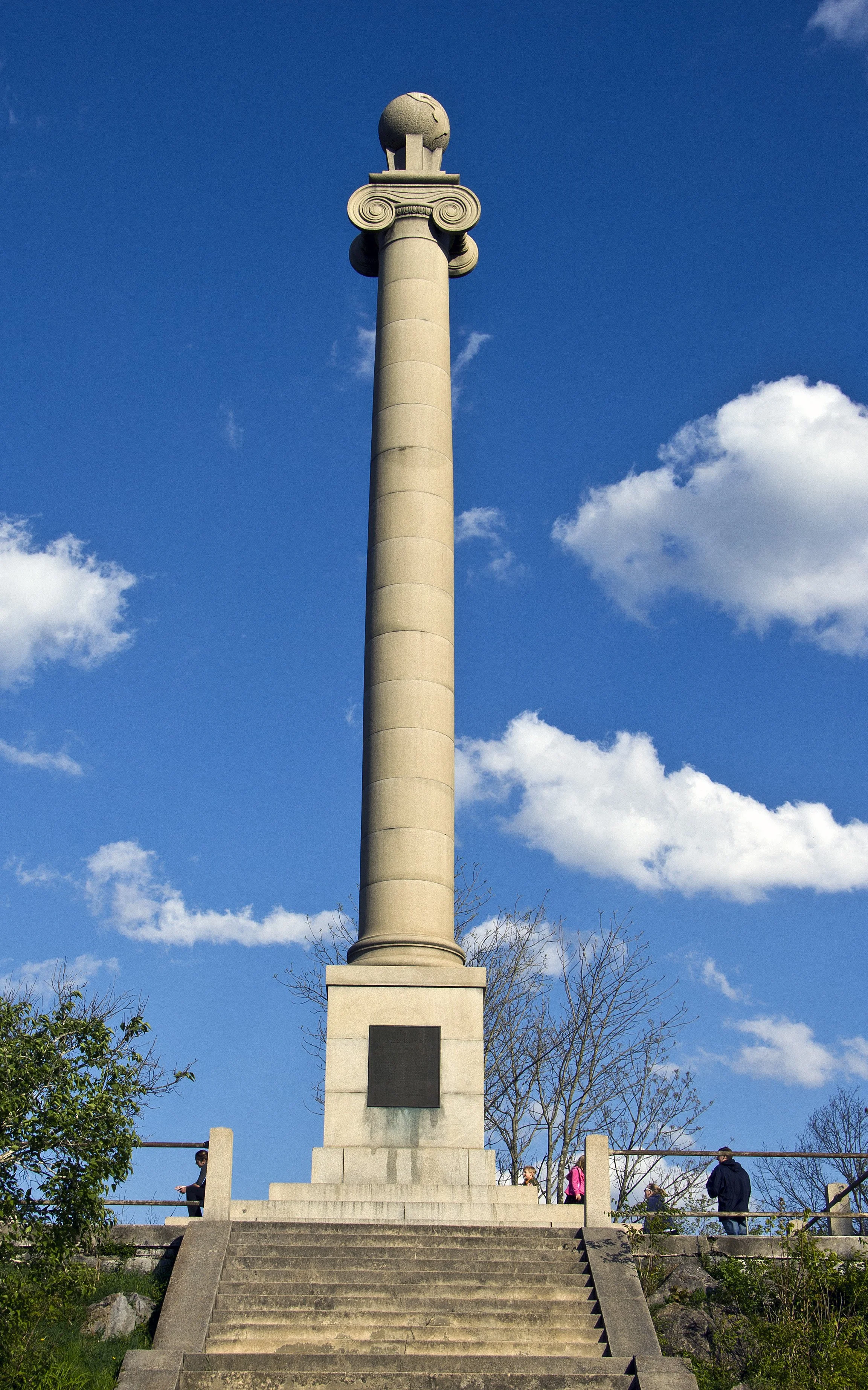 A tall stone monument with a globe on top, set against a blue sky with some clouds. There are steps leading up to the monument and a few people walking nearby.