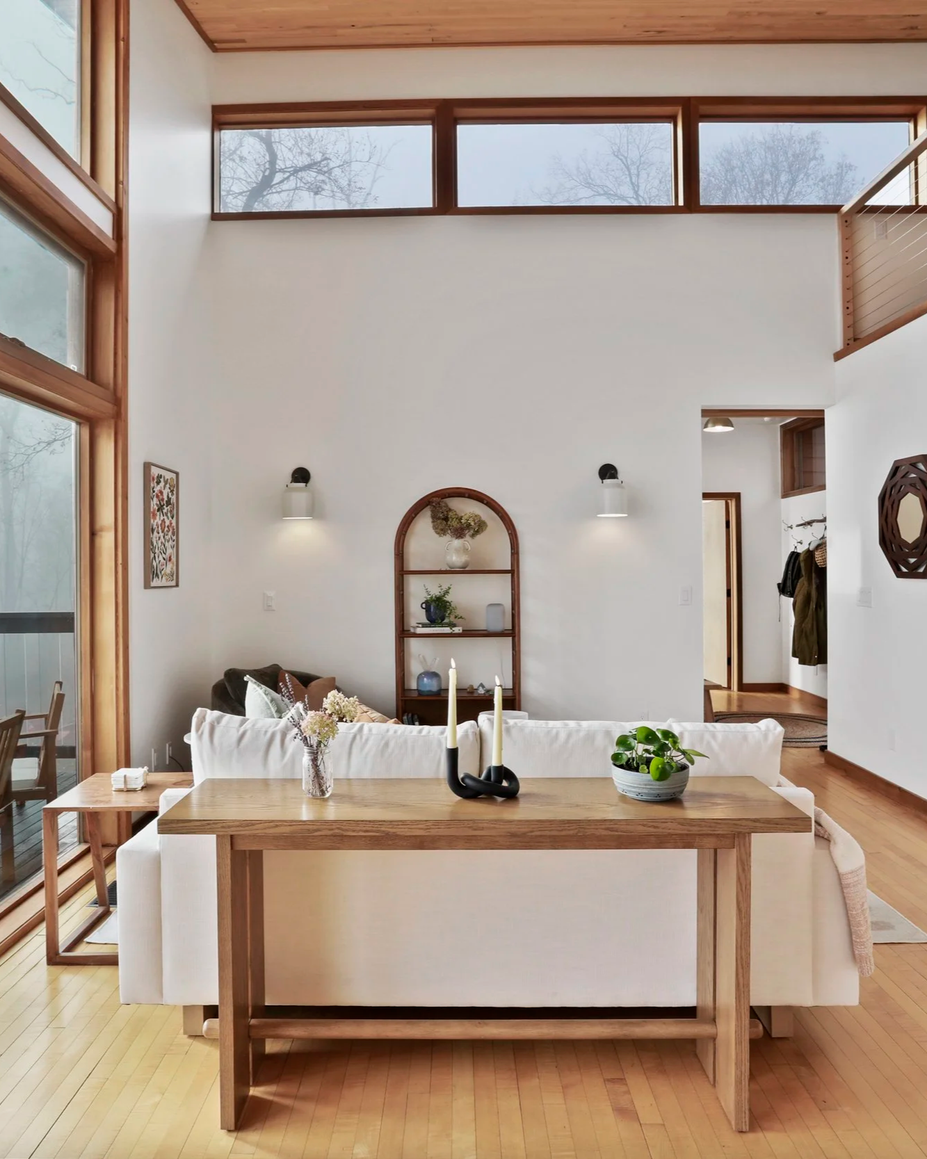 Bright living room with high vaulted ceiling, large windows, white walls, wooden floors, a white sofa, a wooden dining table, and decorative elements such as candles in a black holder, potted plant, and wall art.